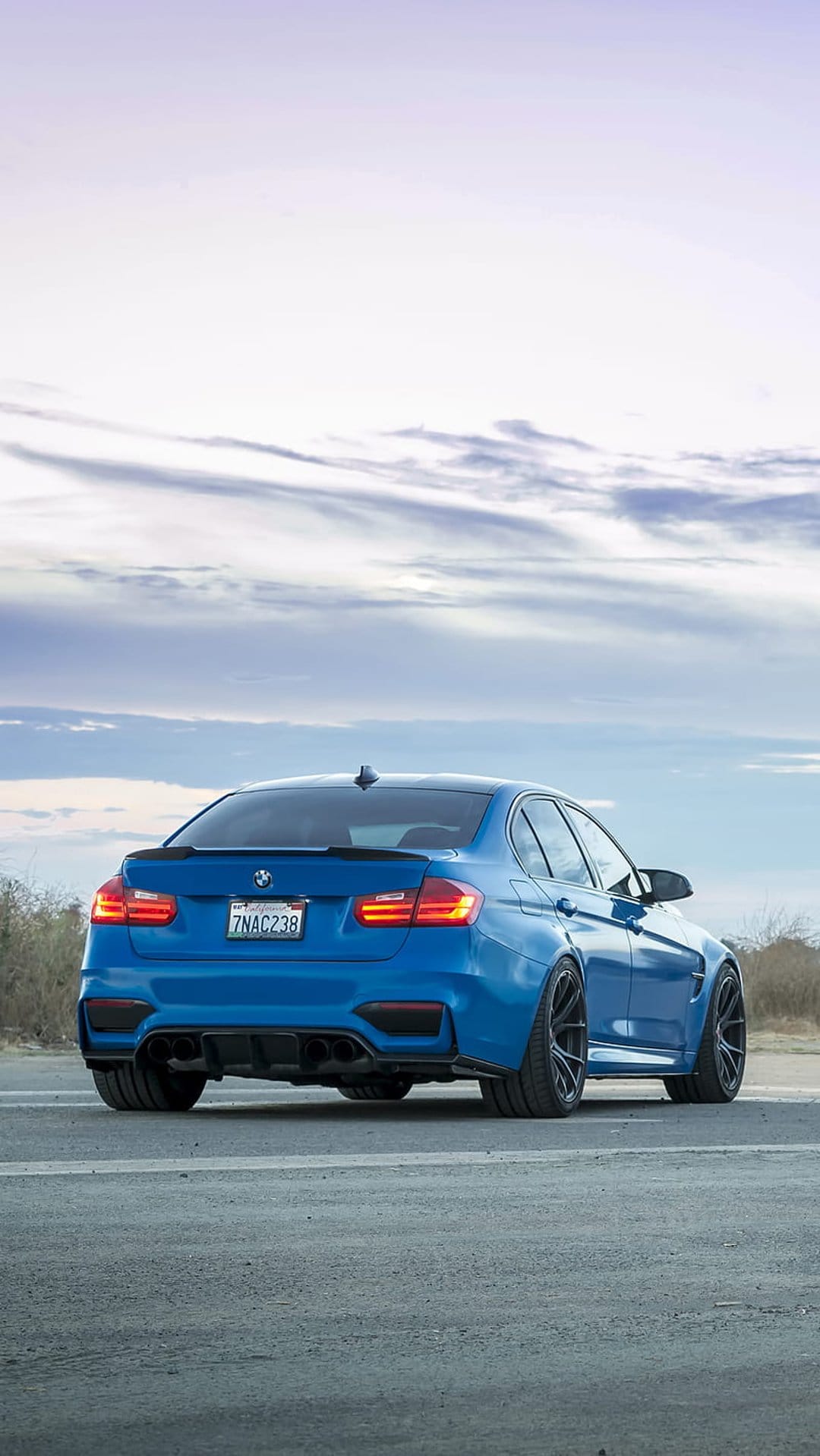 Blue BMW M3 sedan rear view showing quad exhausts on an asphalt road under a cloudy sky during the day.