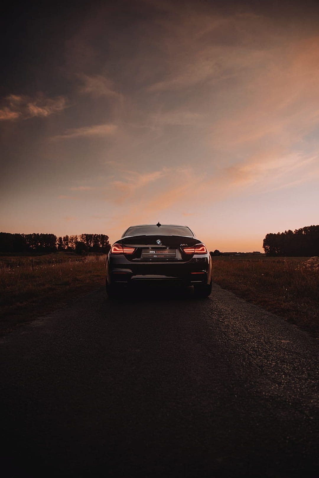 BMW M4 sports car parked on wet asphalt at sunset with glowing taillights and a colorful cloud-filled sky.