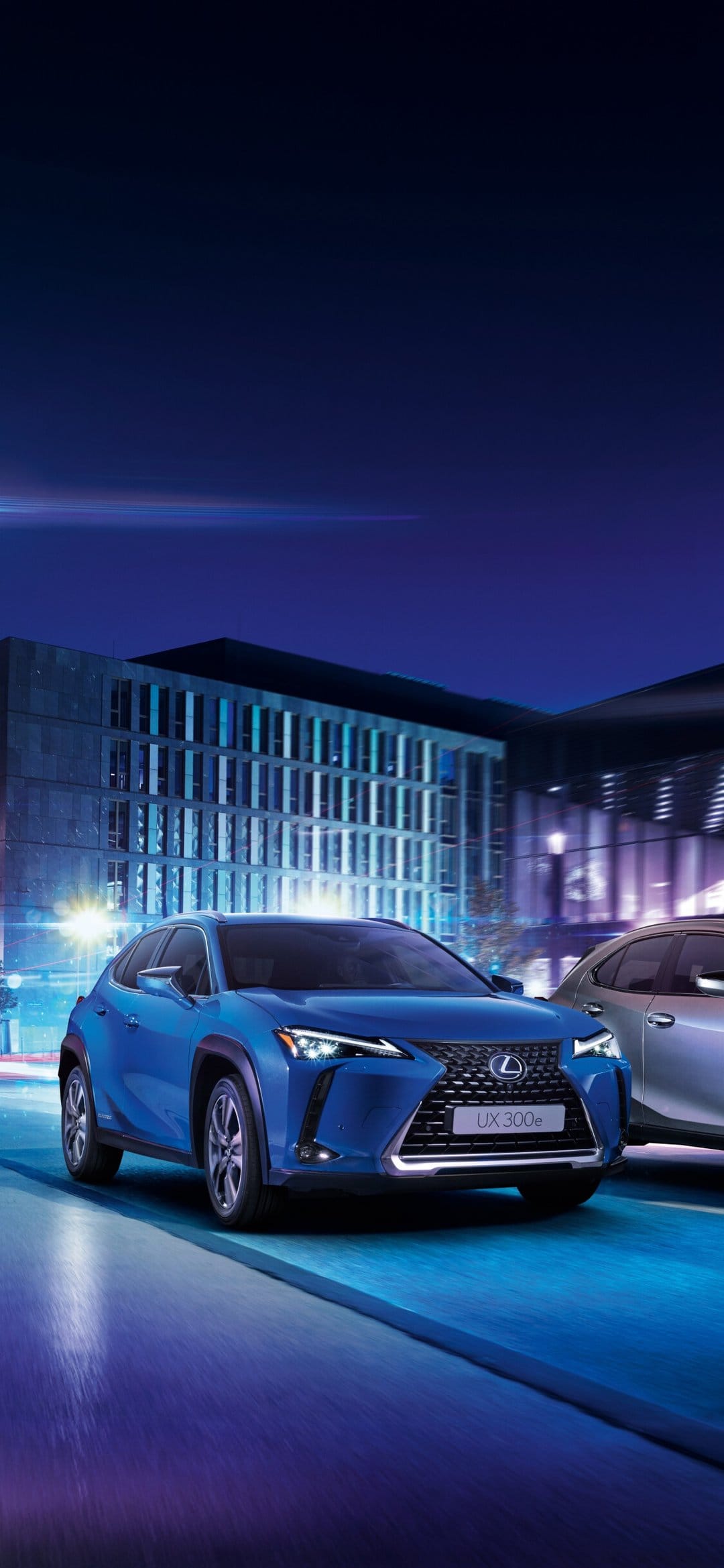Blue Lexus UX 300e electric SUV driving on a city street at night with motion blur and architectural lights.