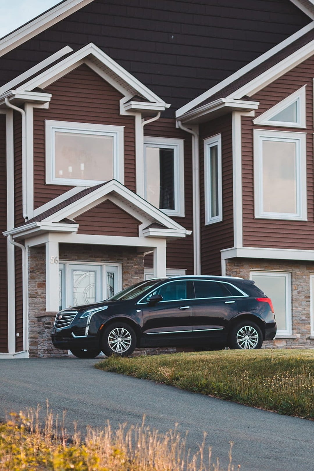 Dark SUV on a paved driveway before a modern house with brown siding, stone masonry, and white gabled roofs.
