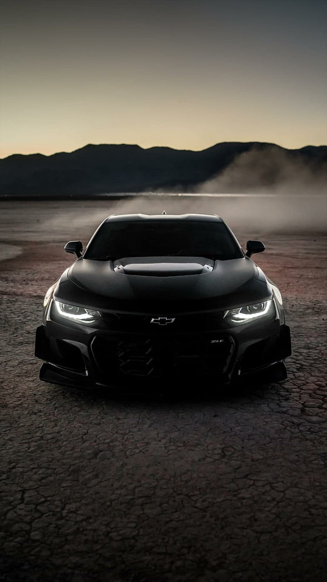 Black Chevrolet Camaro ZL1 with glowing headlights on cracked desert soil under a hazy mountain twilight sky.