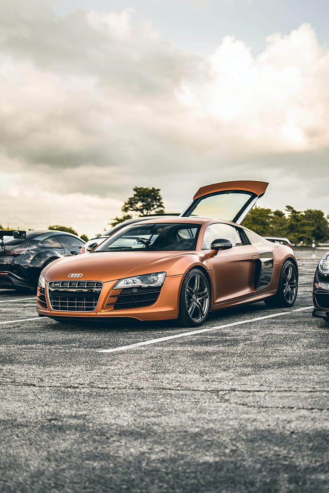 Orange Audi R8 Spyder with open doors parked on asphalt under a cloudy sky during an outdoor car display.
