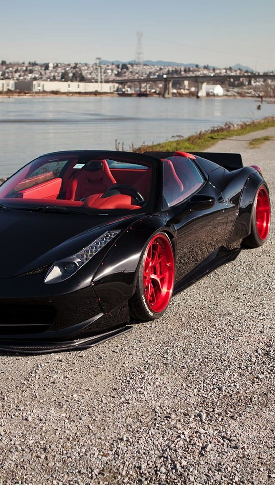 Black Ferrari convertible with red interior and rims parked by water, featuring a bridge and city skyline.
