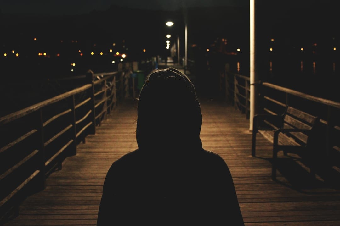 Hooded figure silhouetted on a wooden pier against a distant skyline of glowing city lights and dark water.