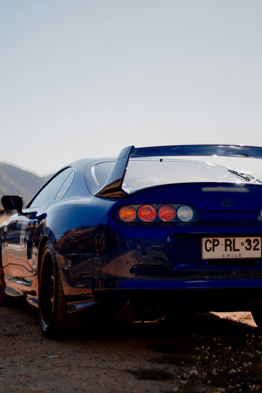 Blue Toyota Supra rear view with round taillights and a large wing parked on a dirt road under a clear sky.