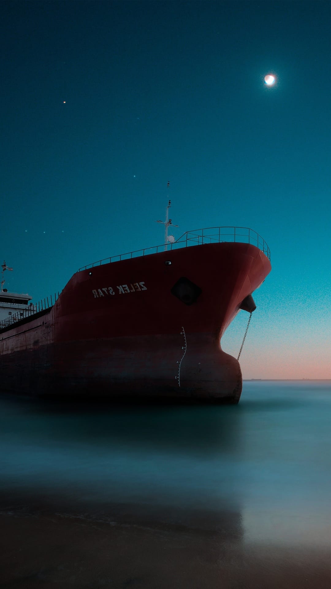 Red cargo ship resting on a dark moonlit beach with a glowing lunar reflection on the calm water surface.