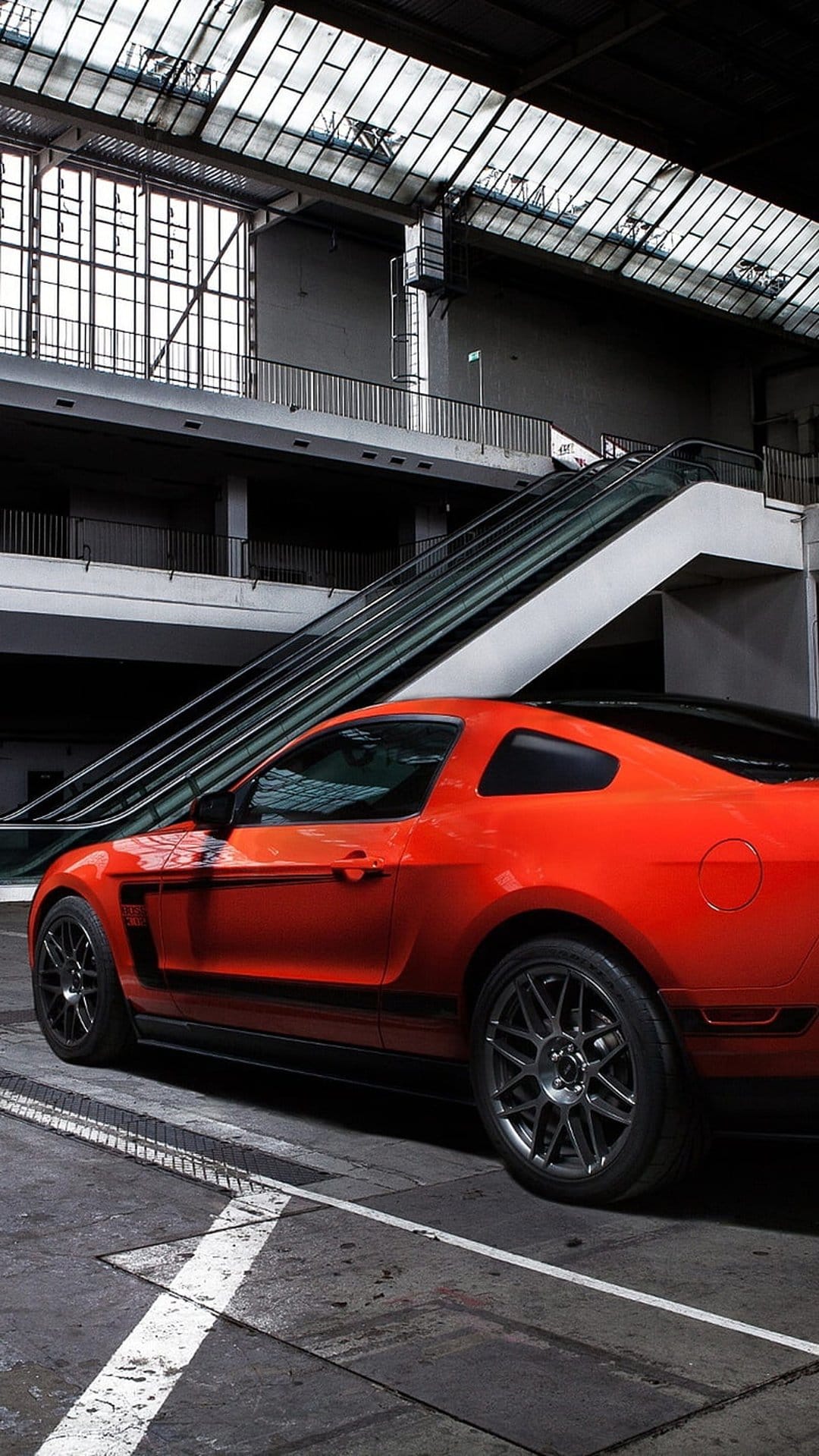 Orange Ford Mustang muscle car sits on a metal escalator inside a modern glass building with industrial lighting.