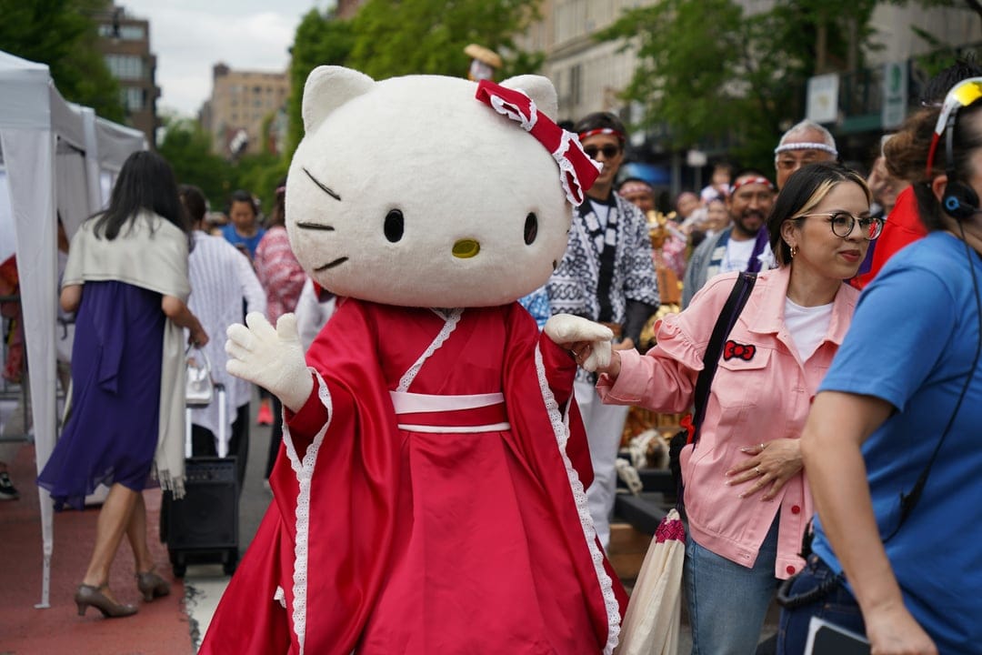 Hello Kitty mascot in a red kimono stands before a crowded street festival with white tents and a diverse parade.
