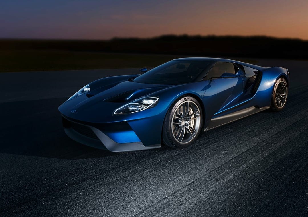 Blue Ford GT supercar on a dark track at sunset with glowing headlights and a colorful twilight sky background.