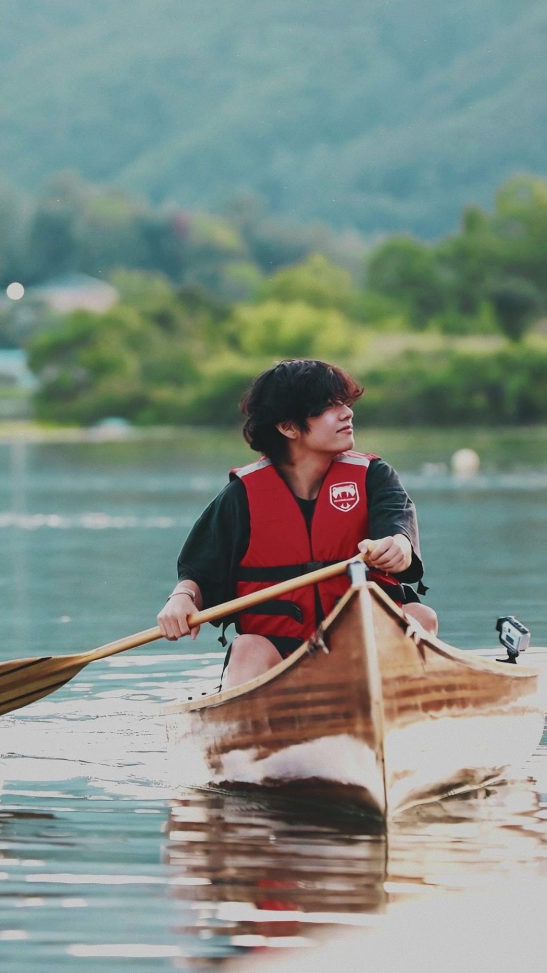 Man in a red life vest rows a wooden canoe on a glassy lake surrounded by lush green hills and soft sunlight.