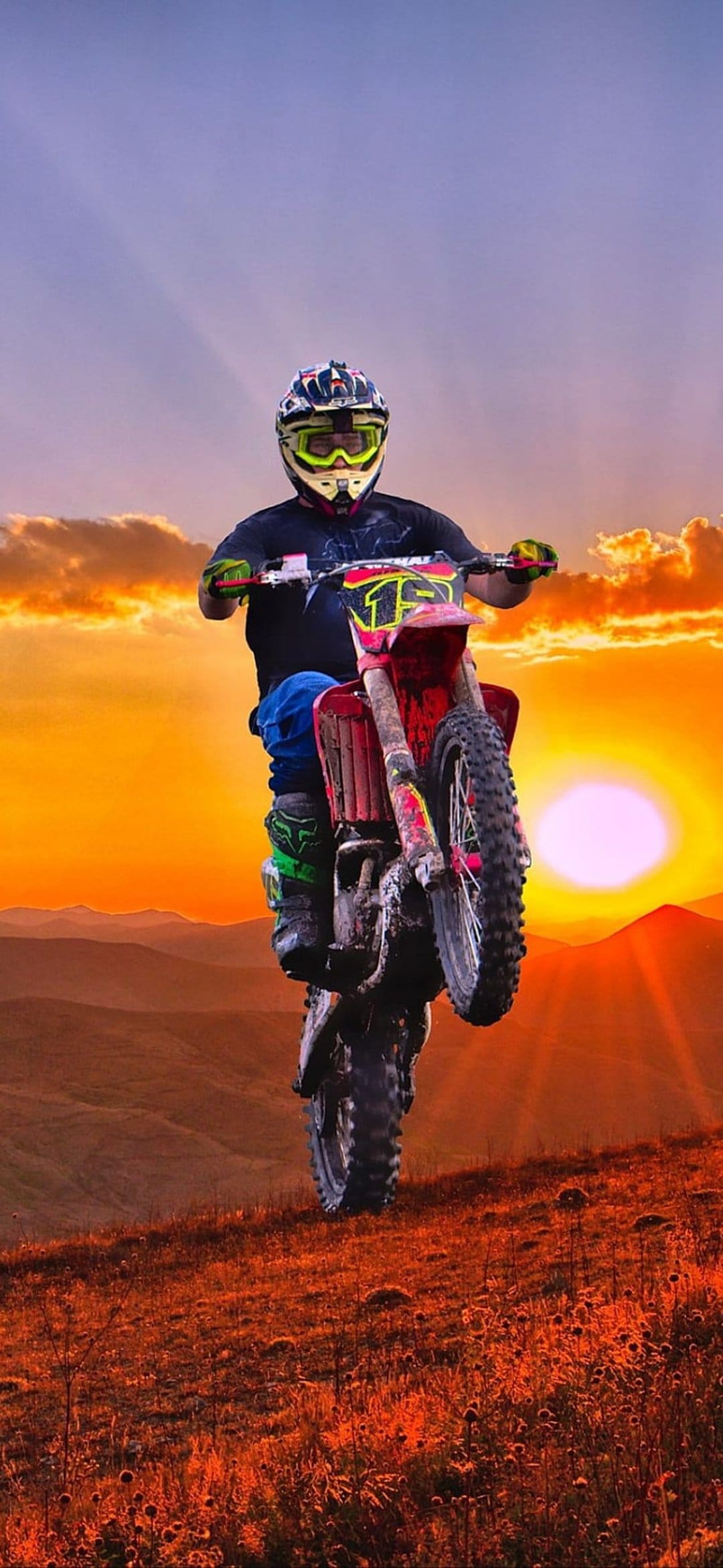 Motocross rider on a red dirt bike performs a wheelie on a hill under a purple and orange sunset sky.