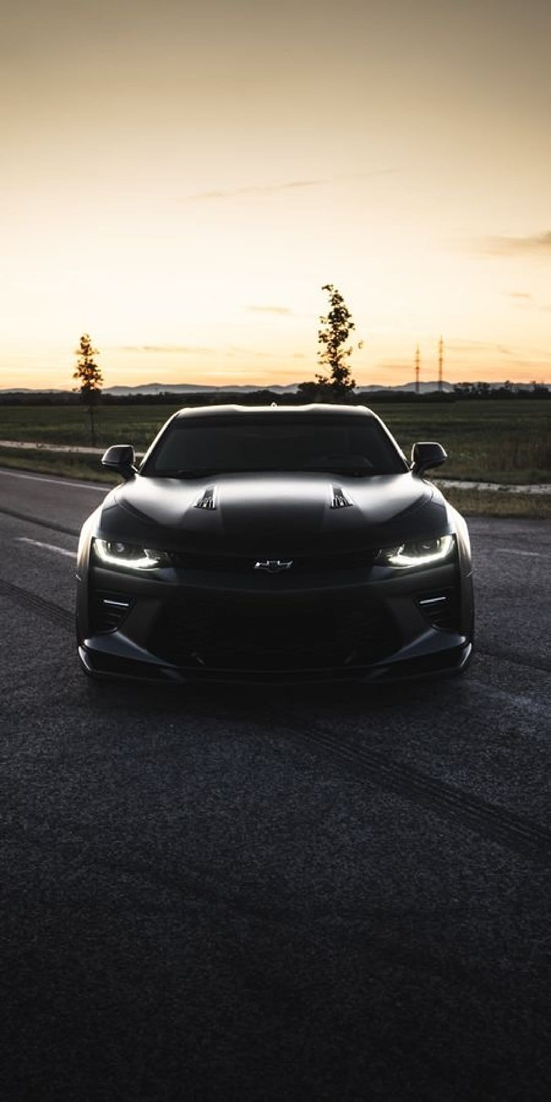 Black Chevrolet Camaro with glowing headlights parked on an asphalt road during a vibrant orange sunset.