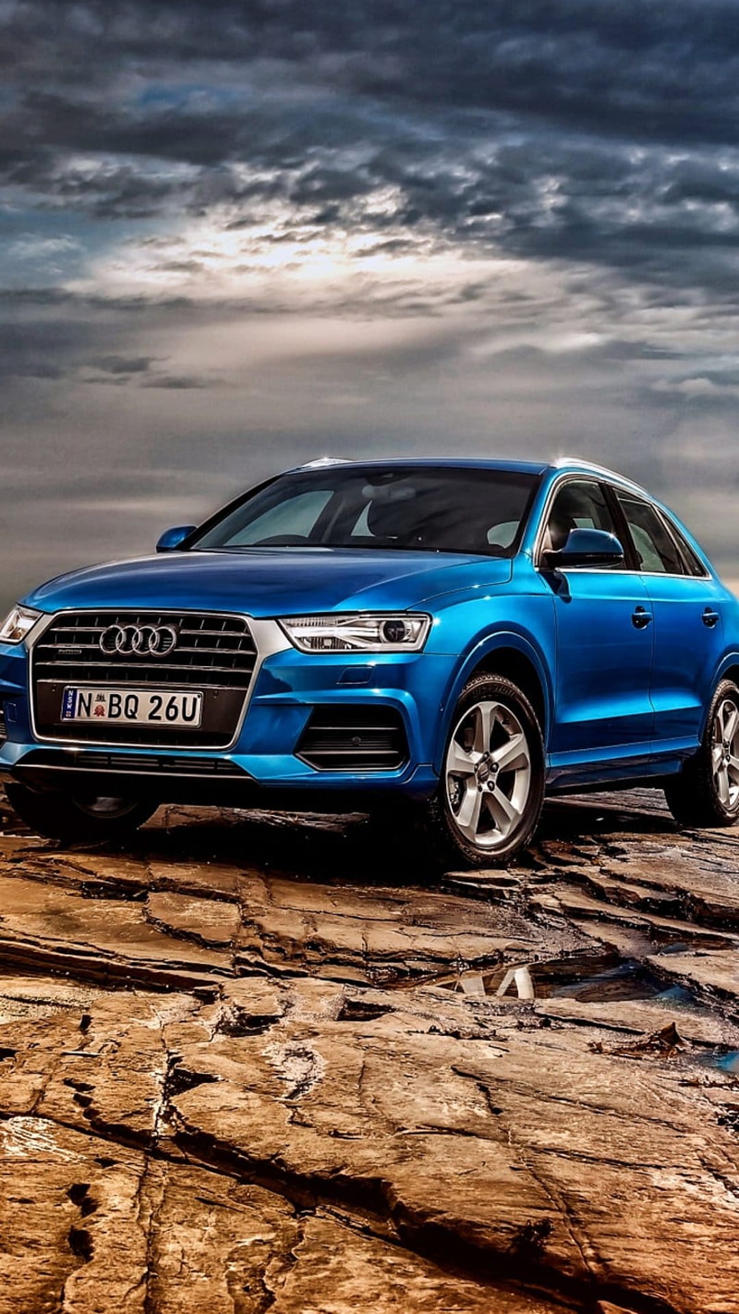 Blue Audi Q3 SUV parked on dark textured rocks beneath a dramatic overcast sky in a vertical outdoor scene.