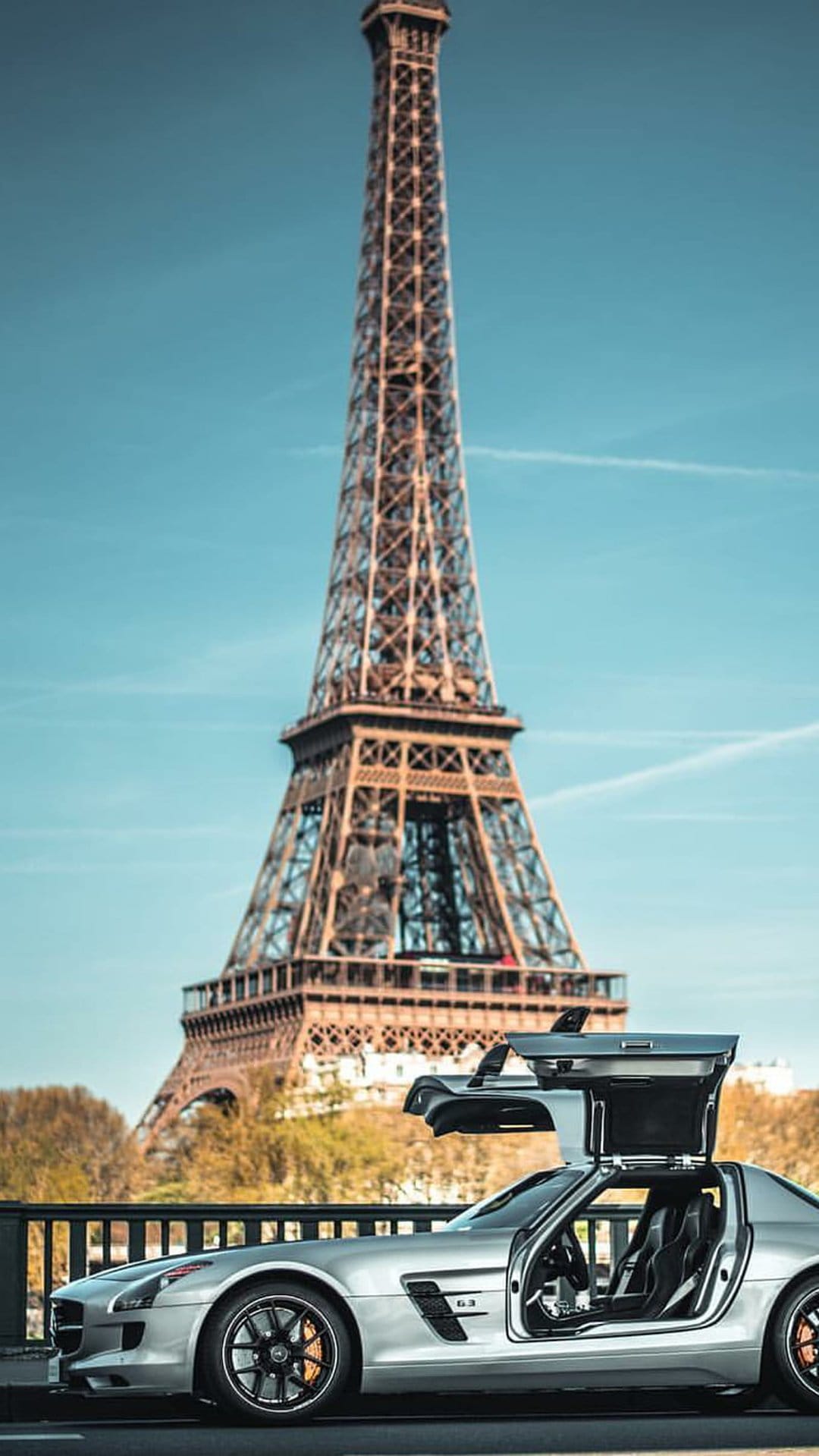 Silver Mercedes SLS AMG with open gullwing doors parked on a Paris bridge with the Eiffel Tower in the background.