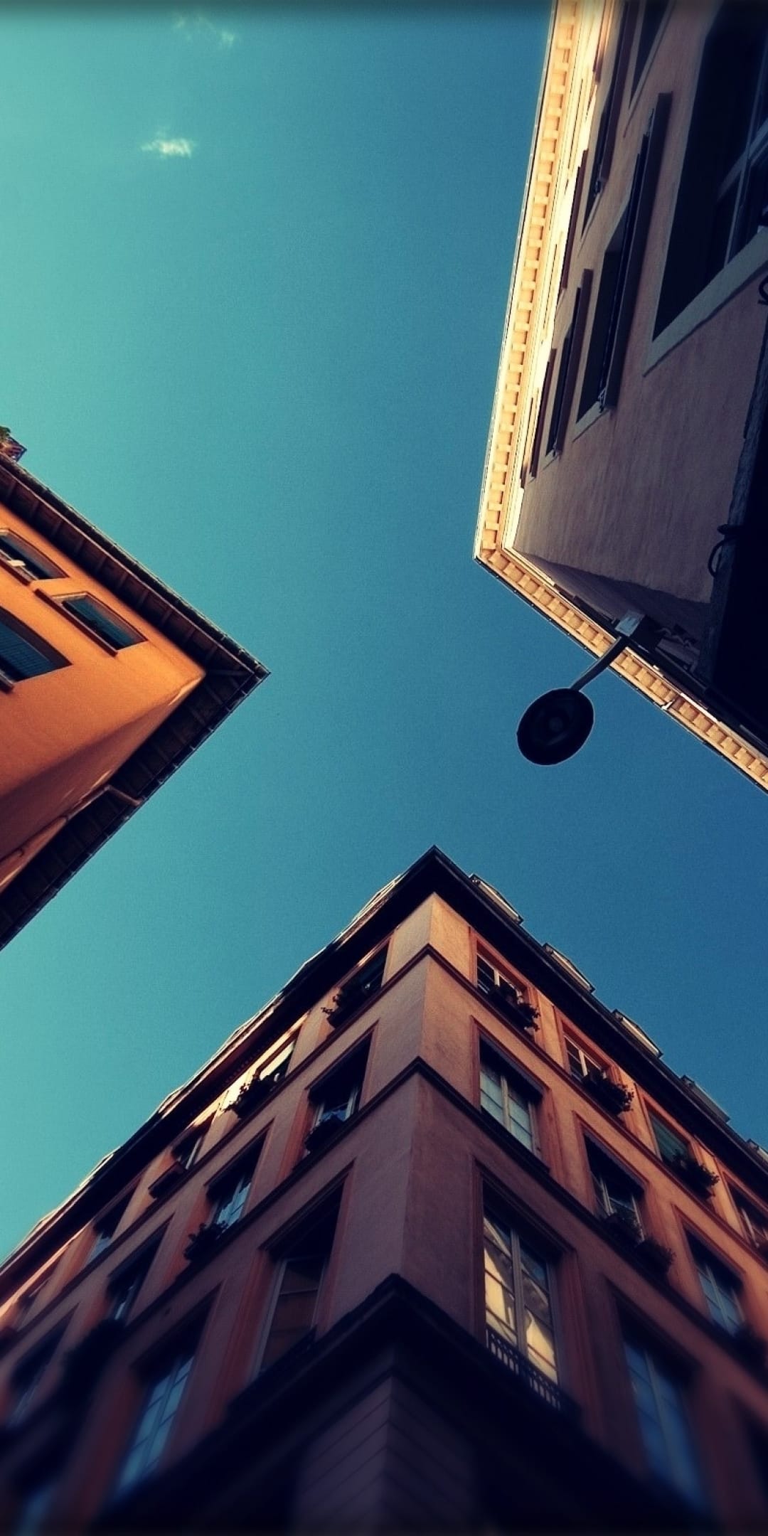 Three warm-toned buildings angle upward toward a clear blue sky with a silhouetted streetlamp in the foreground.