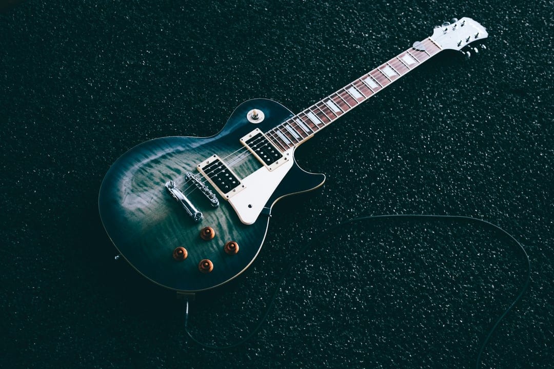 Teal electric guitar with a flame maple top, white pickguard, and coiled cable on a black textured surface.