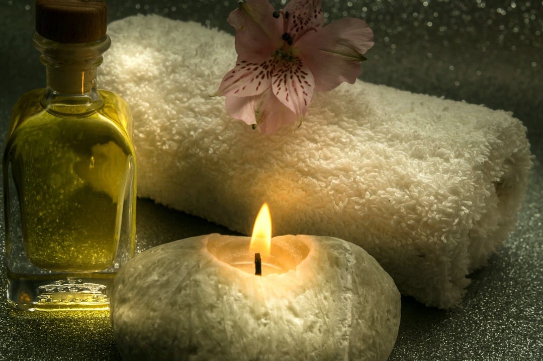 Lit candle in a stone holder beside golden oil, a folded white towel, and a soft pink flower on a flat surface.