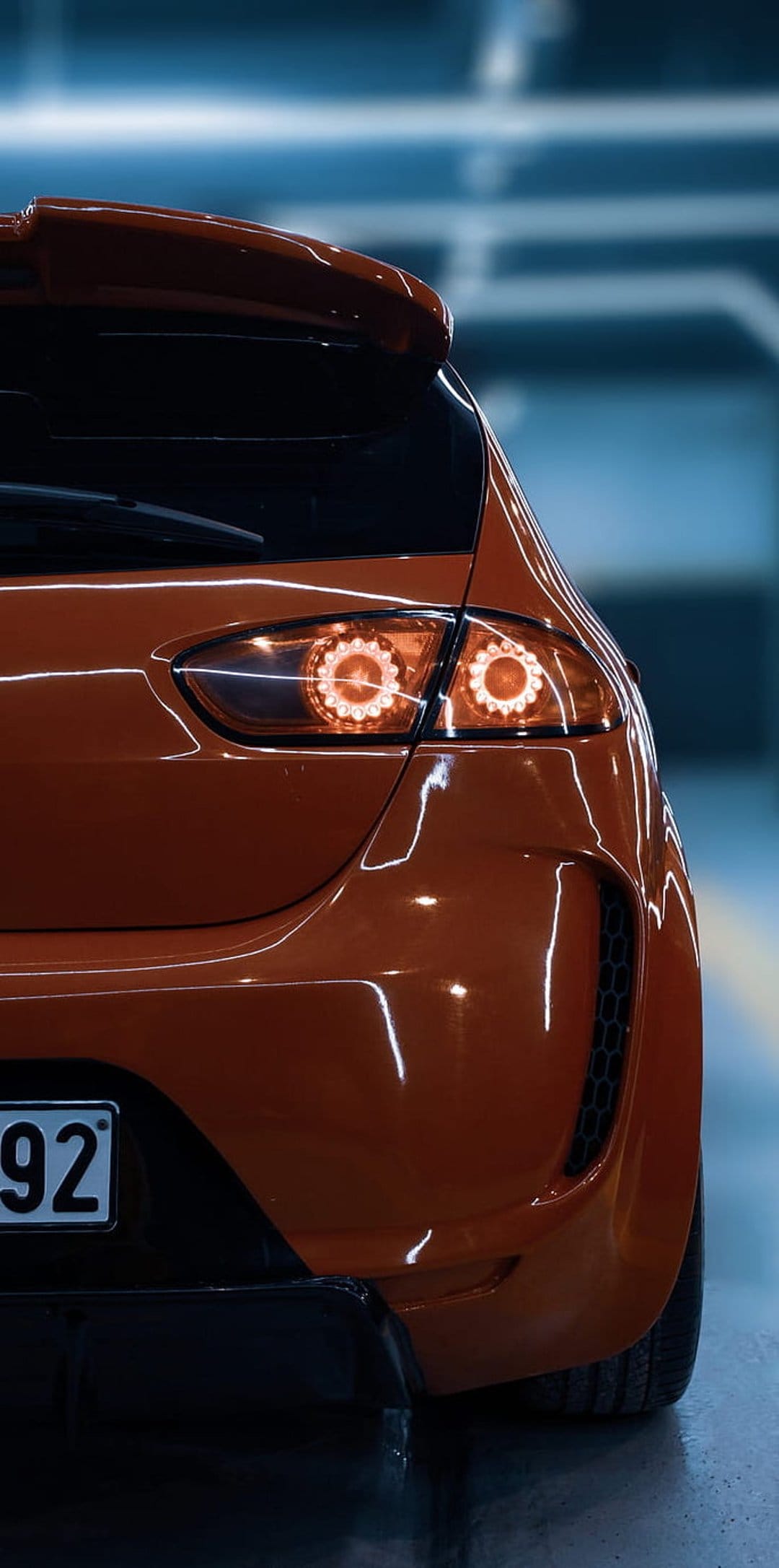 Orange sports car rear taillights and spoiler glowing with warm light on a glossy metallic vehicle surface.