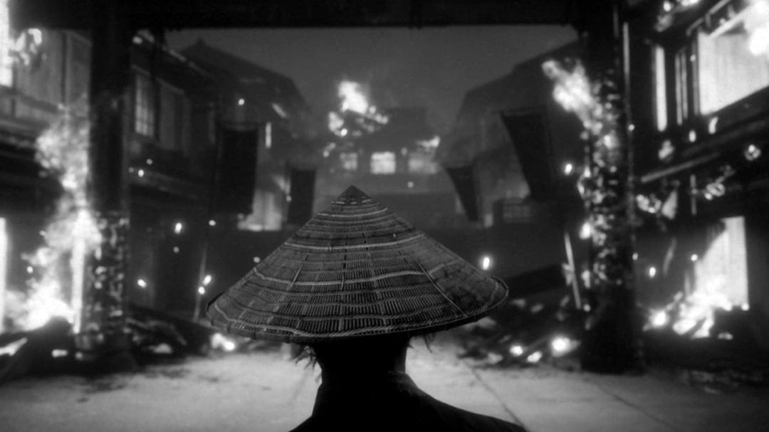 Rear view of a person in a conical hat looking at a burning historical Japanese village in black and white.