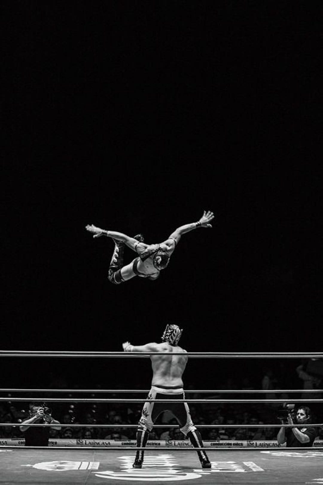 Masked wrestler suspended mid-air above a dark wrestling ring during a high-flying acrobatic maneuver.