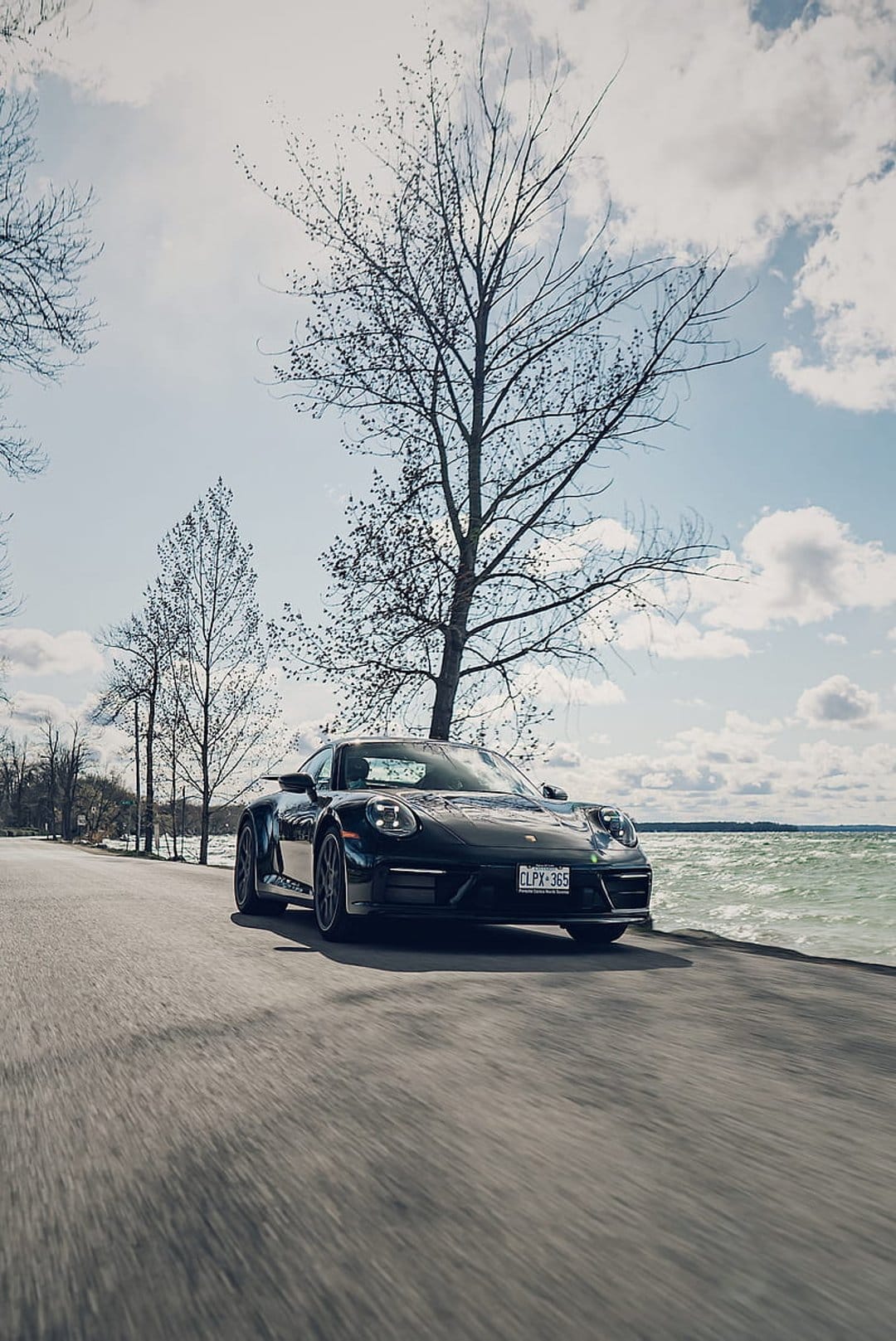 Black Porsche 911 sports car drives along a scenic ocean highway with motion blur and bare trees.