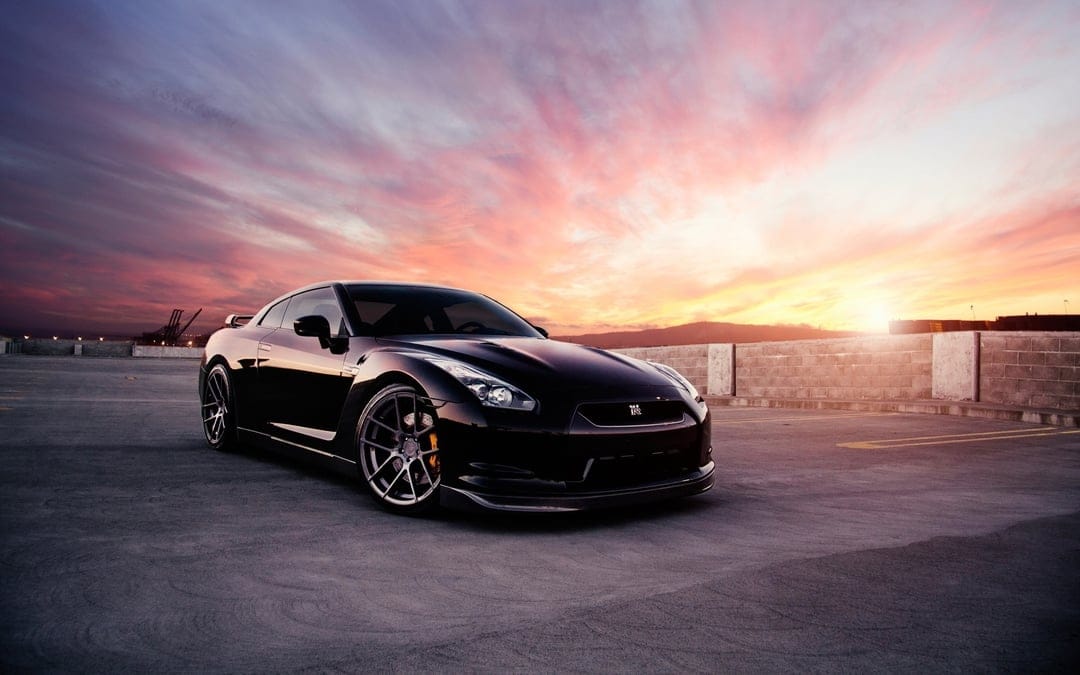 Black Nissan GT-R parked on a concrete lot under a pink and orange sunset sky with wispy clouds.
