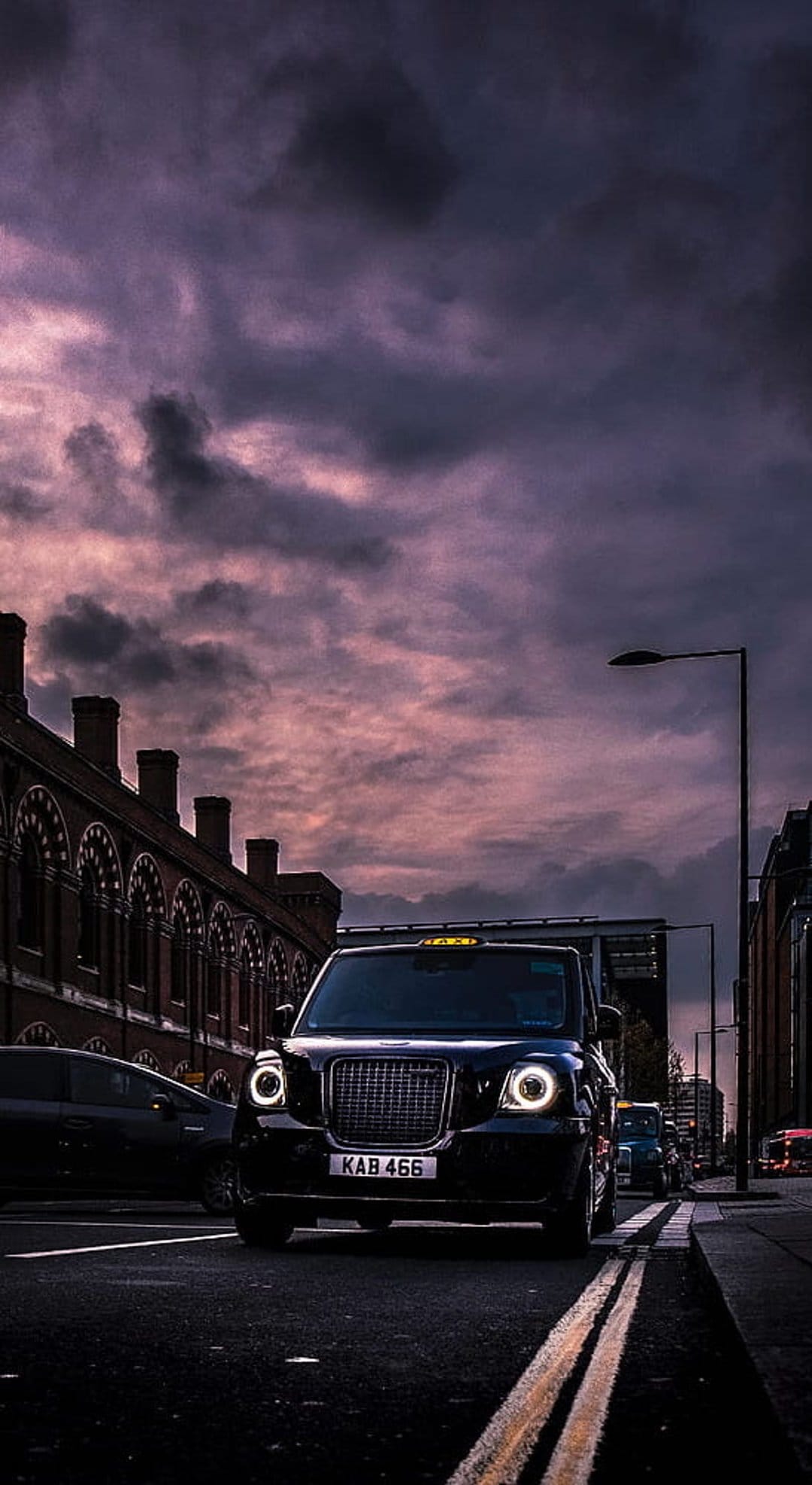 Black London taxi cab with glowing headlights driving on a city street under a dramatic purple and grey dusk sky.