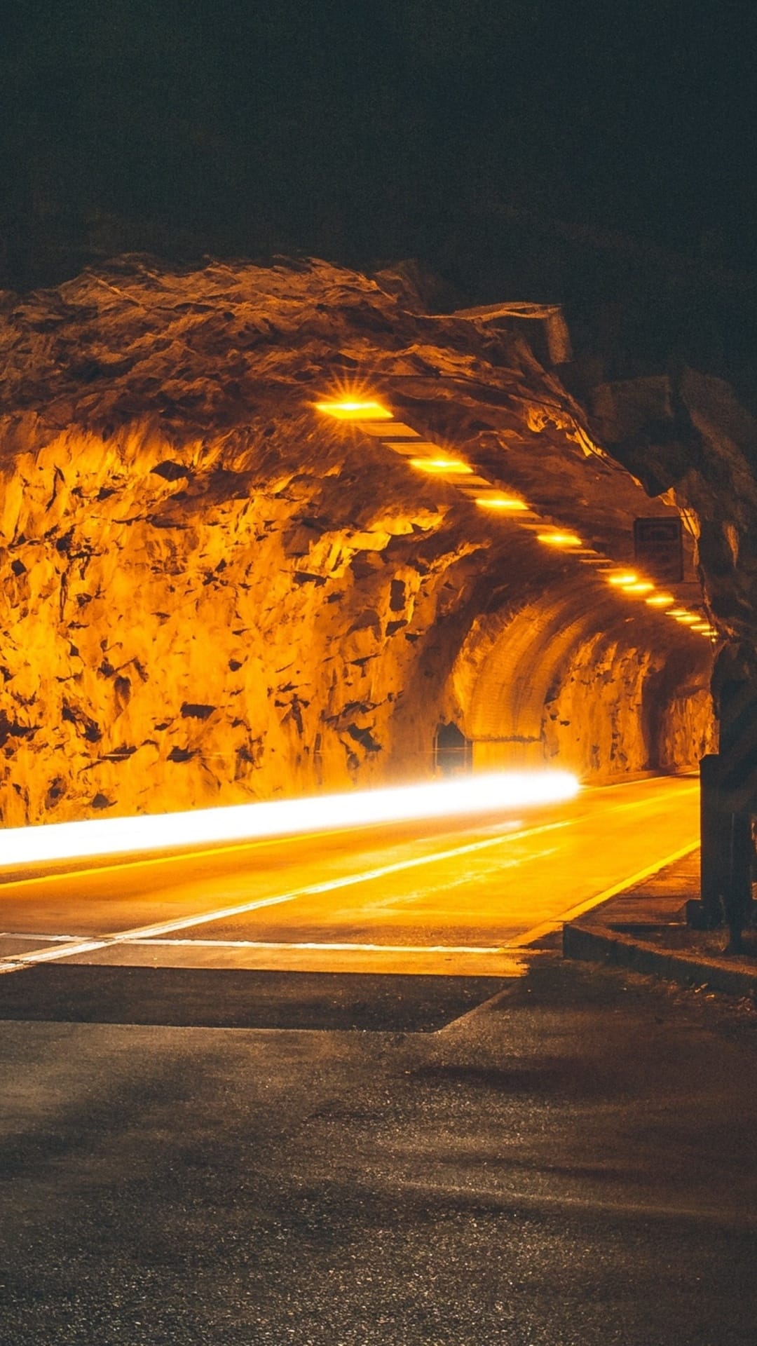 Amber car light trails streak through a jagged rock-hewn tunnel illuminated by overhead industrial lamps.