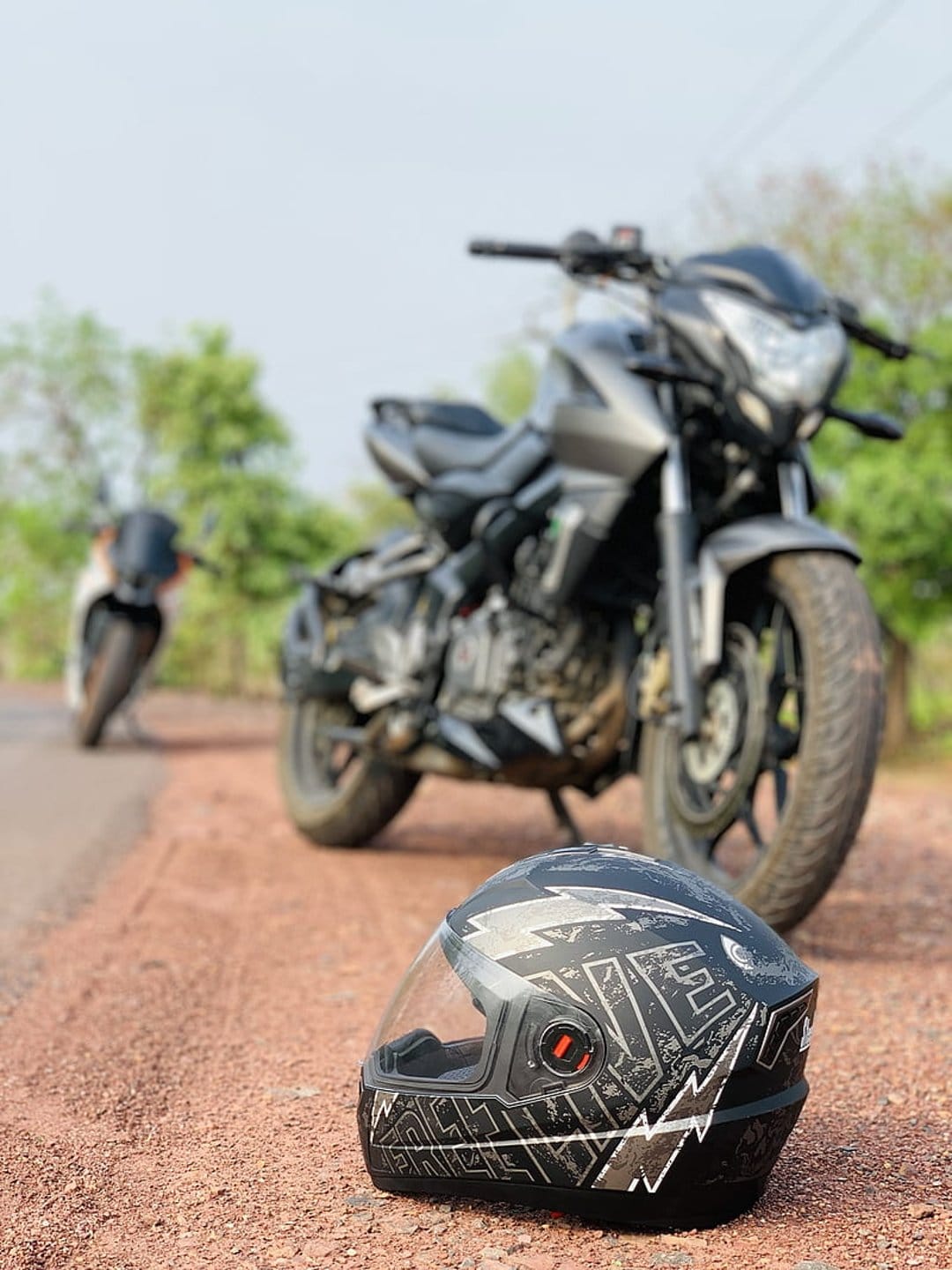Black motorcycle and full-face helmet with white graphics rest on a dirt road against a blurred natural background.
