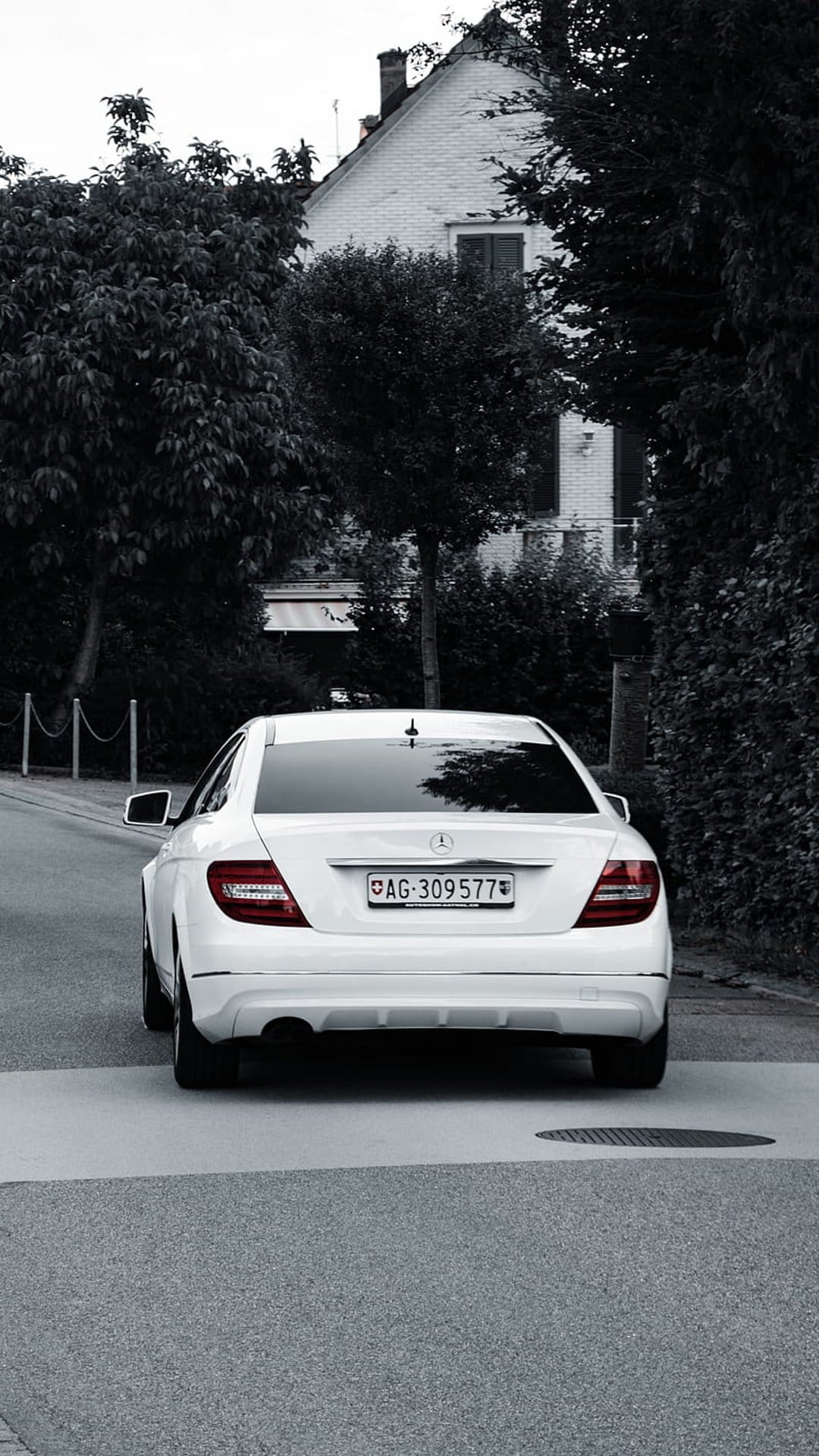White Mercedes-Benz sedan rear view parked on an asphalt driveway near a suburban house and leafy green trees.