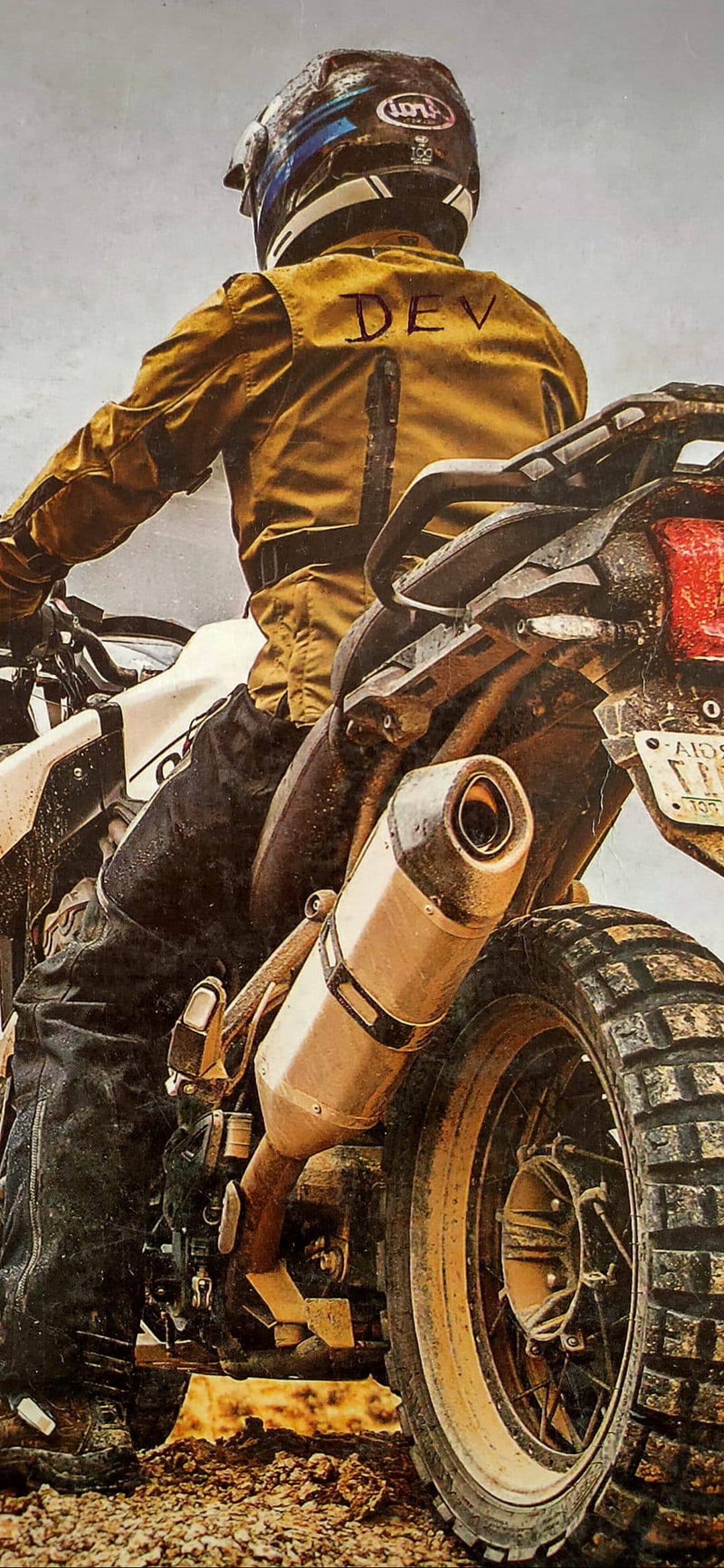 Dirt bike rider wearing a yellow DEV jacket and helmet rides through mud, focusing on the rear tire and exhaust.