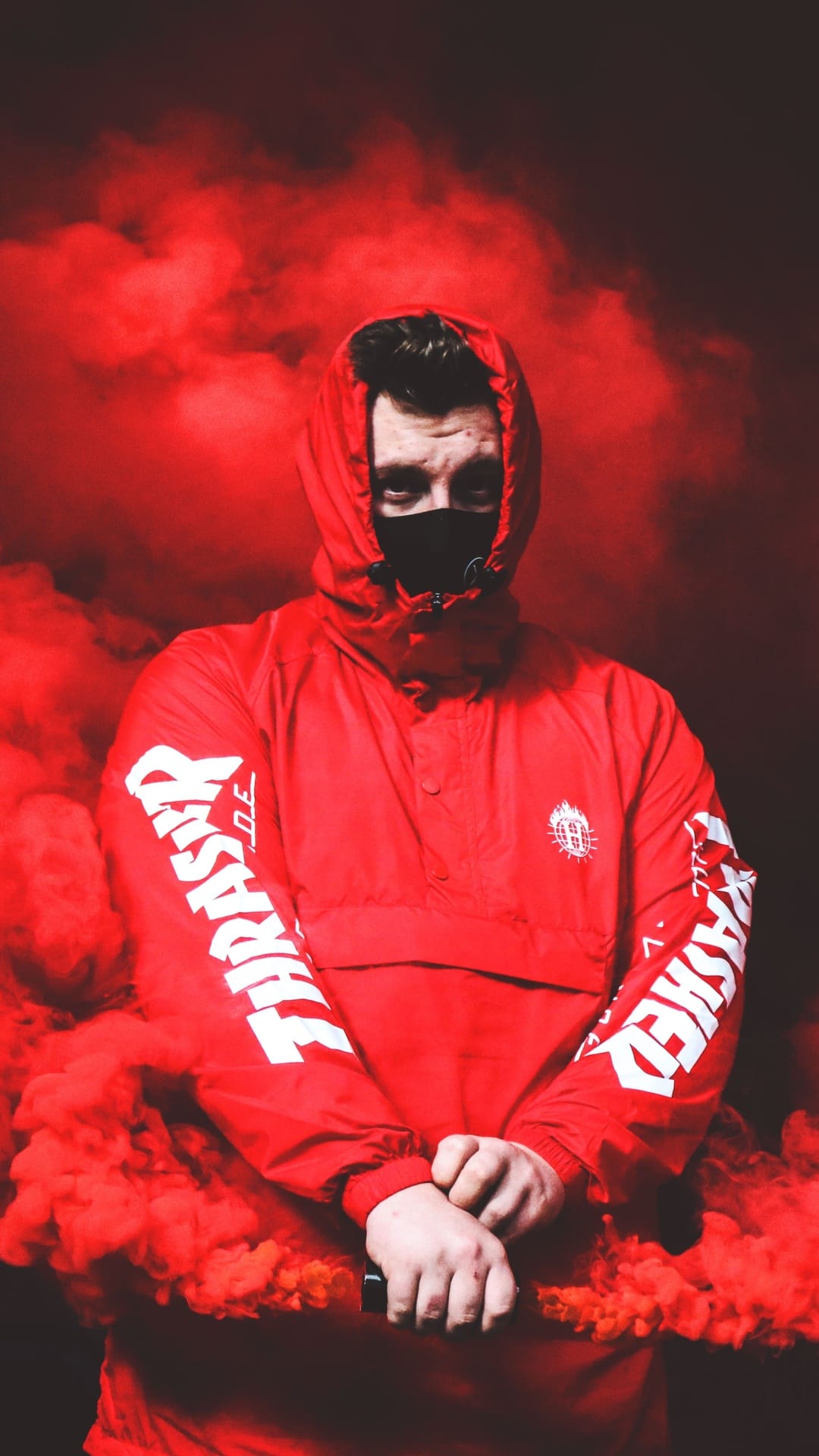 Masked person in a red jacket with white sleeve lettering standing within a dense cloud of crimson smoke.
