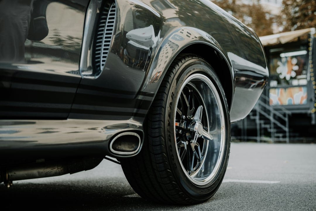 Chrome rim of a dark muscle car with a visible exhaust pipe and tire tread against a blurred urban background.