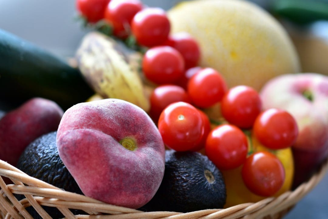 Ripe red tomatoes, a fuzzy peach, and avocados rest in a woven basket alongside a banana and green zucchini.
