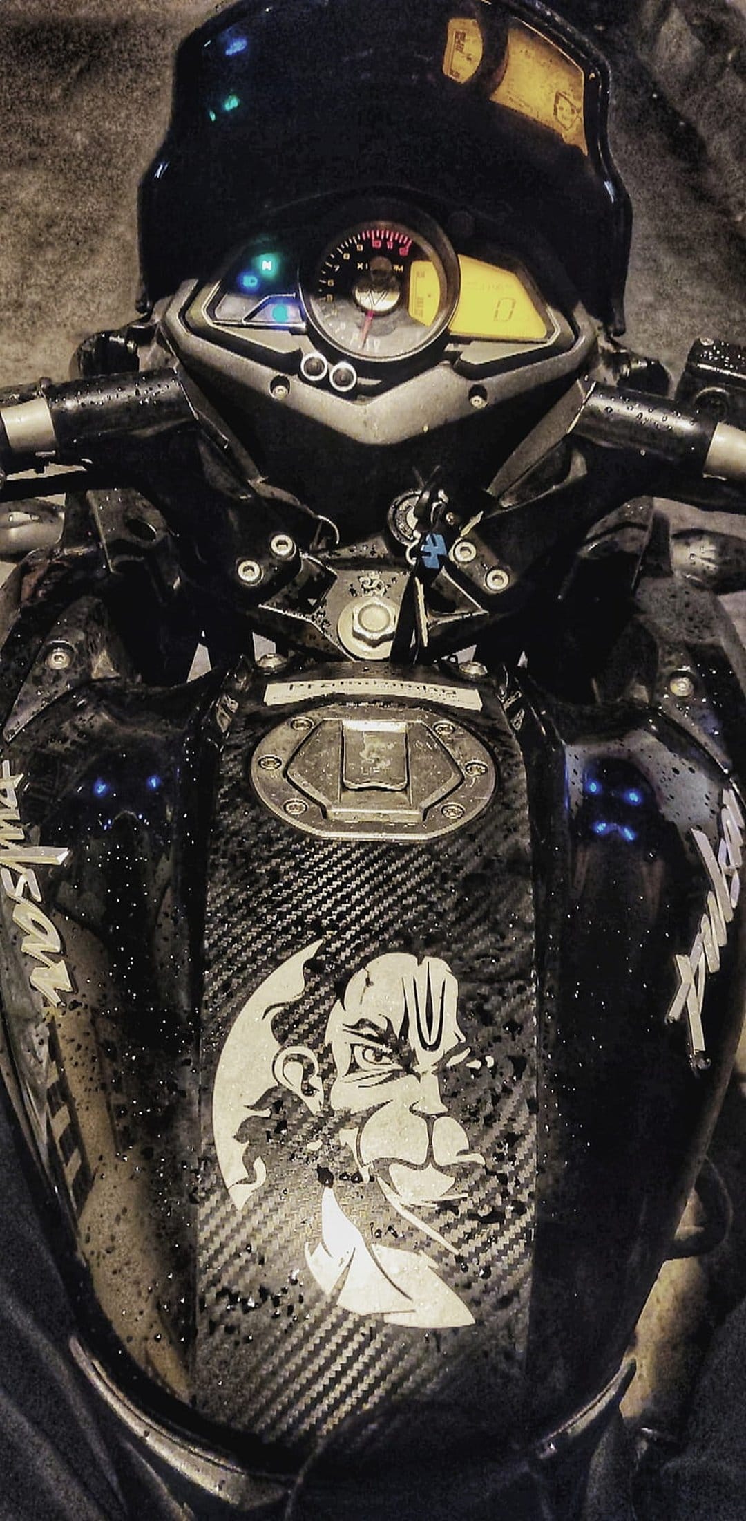 Hanuman sticker on a black carbon fiber motorcycle fuel tank with water droplets and handlebars in the background.