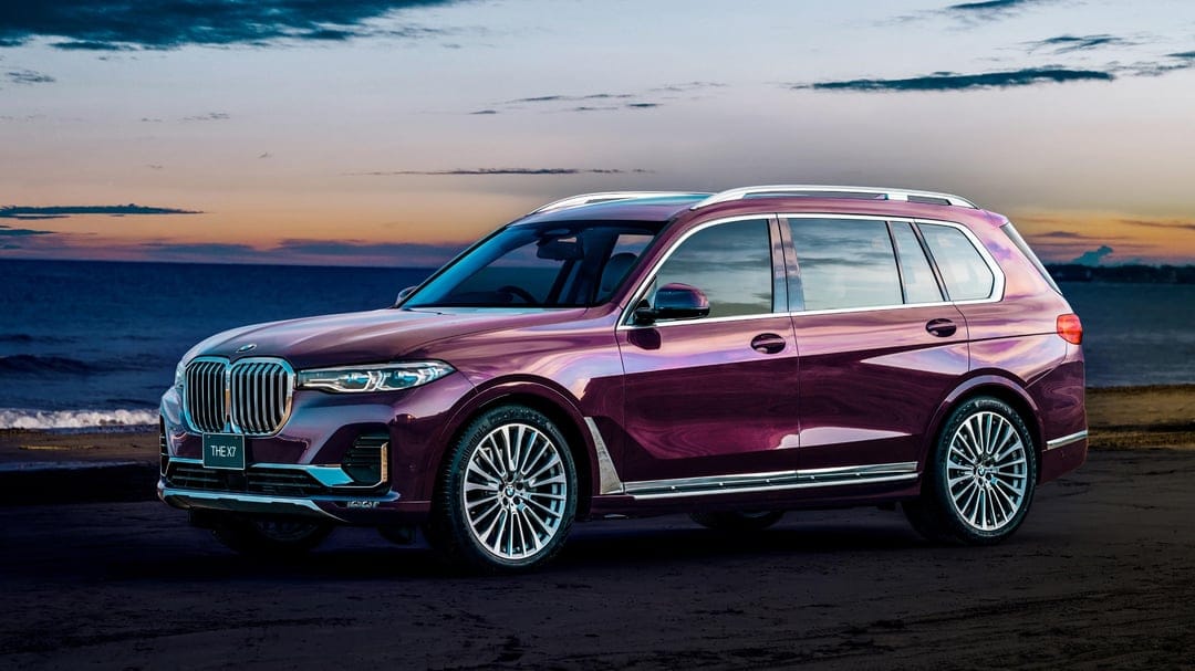 Purple BMW X7 SUV parked on a sandy beach at sunset with a dramatic cloudy sky and ocean waves behind it.