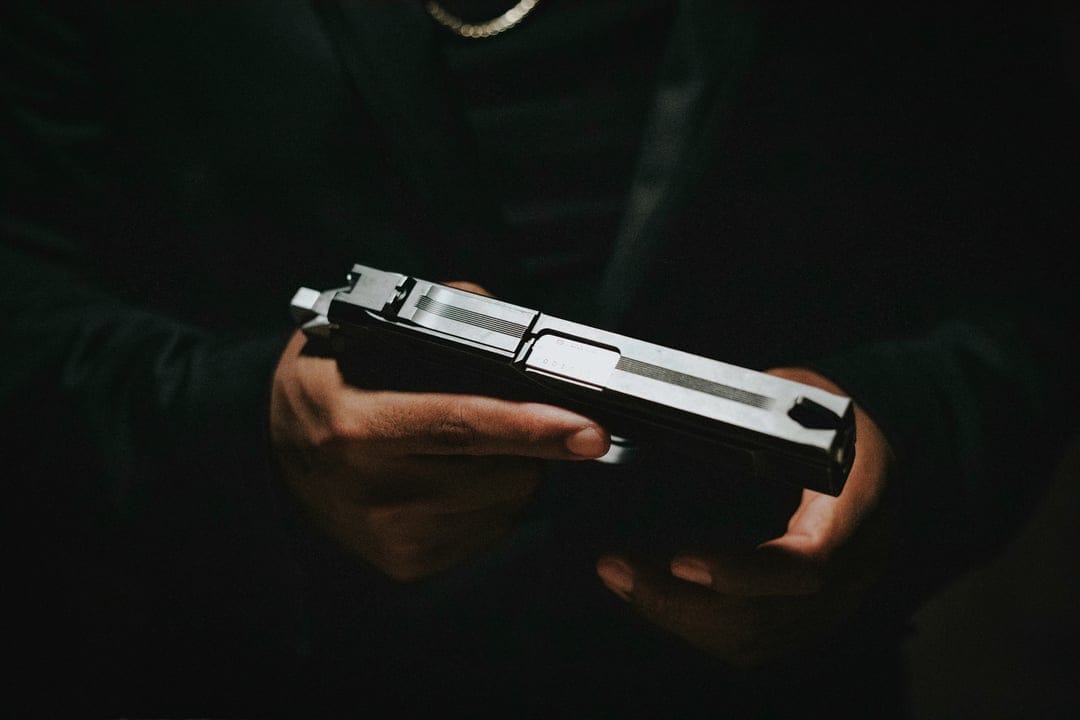 Hands grip a sleek black semi-automatic pistol under harsh dramatic lighting with a dark blurred background.