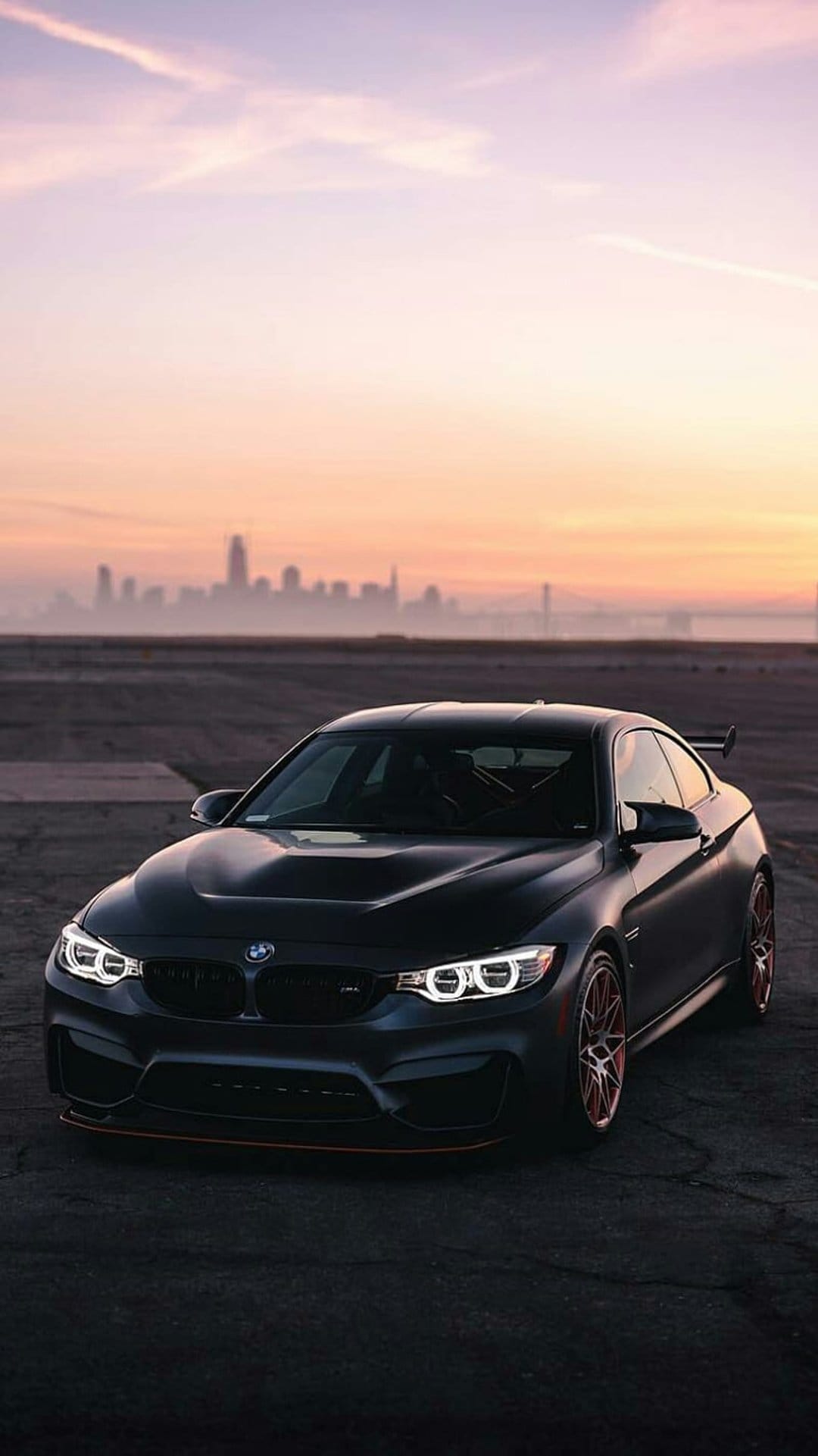 Dark BMW M4 Coupe with red-accented rims parked on asphalt during a hazy orange city skyline sunset at dusk.