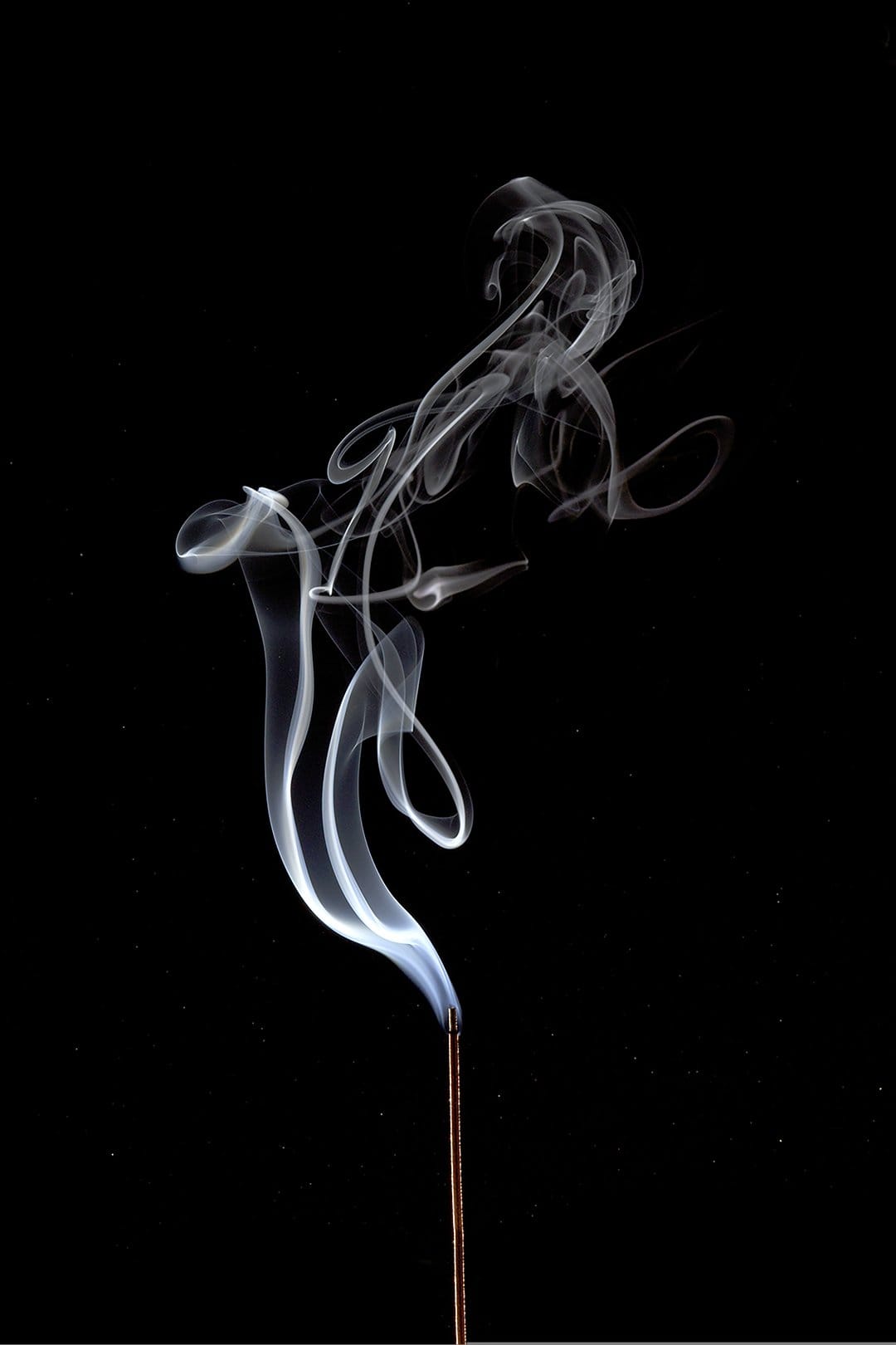 Intricate white incense smoke curls rise in delicate translucent patterns against a deep black backdrop.