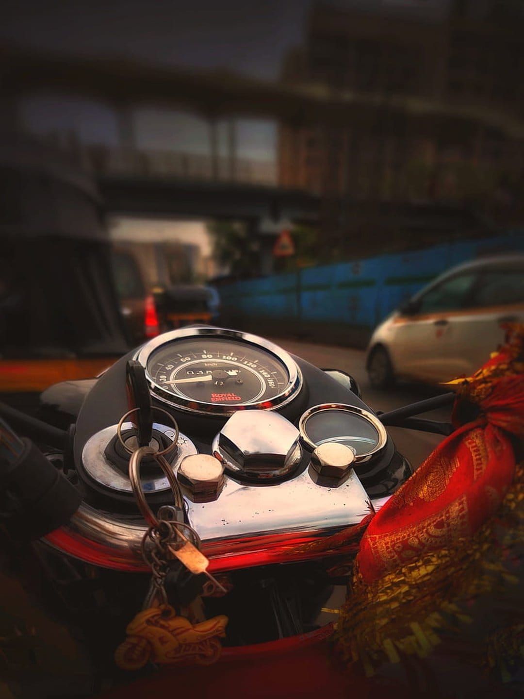 Motorcycle speedometer and ignition keys with a red patterned cloth draped over the handlebars in a city.