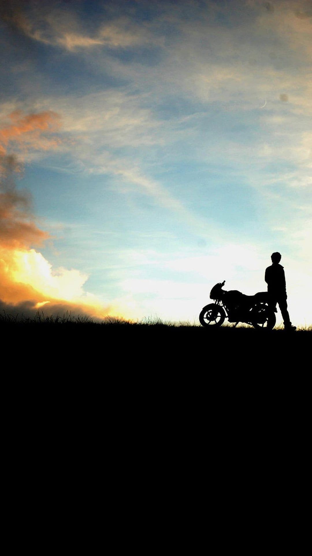 Biker silhouette stands next to a motorcycle under orange and blue twilight clouds in a wide outdoor setting.