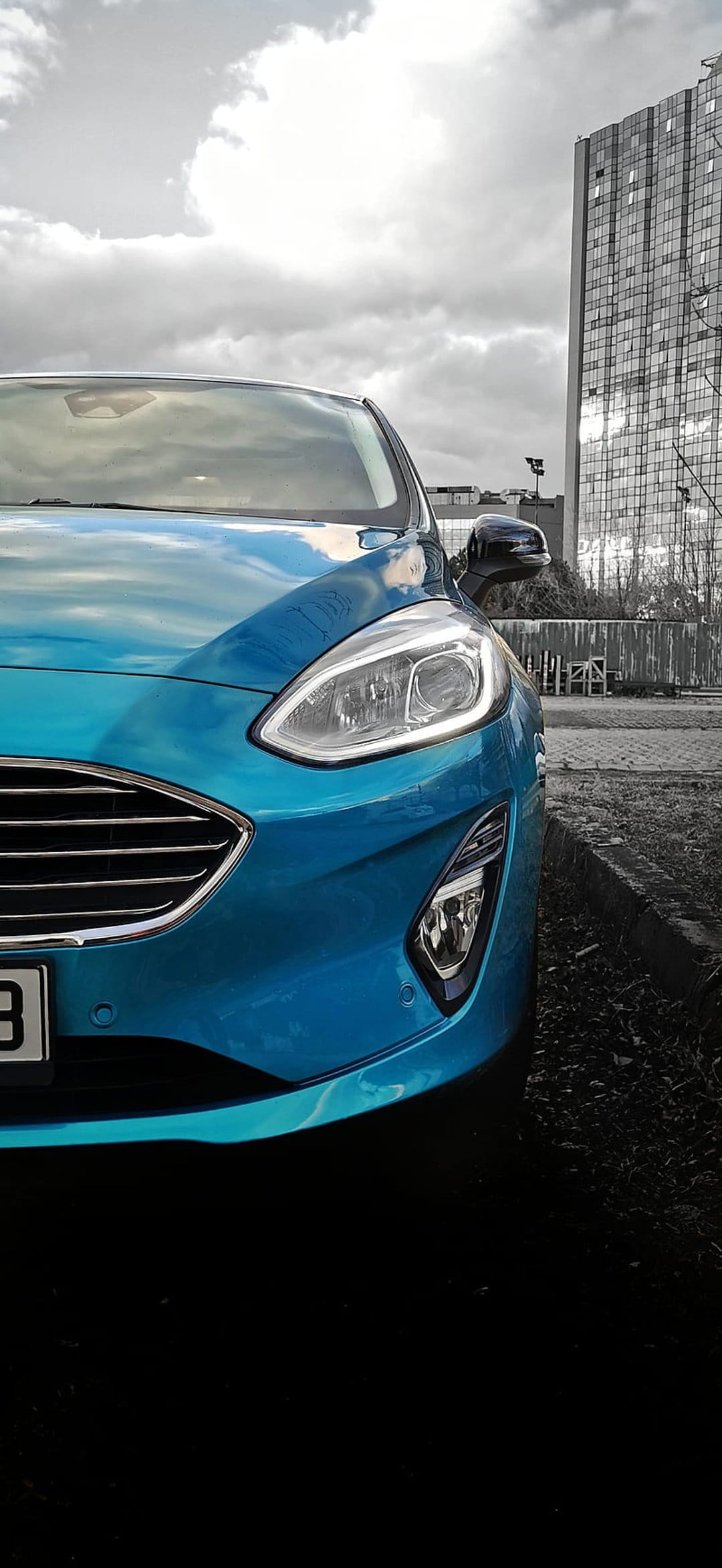 Blue Ford Fiesta parked at a low angle in front of a modern glass skyscraper under a cloudy grey sky.