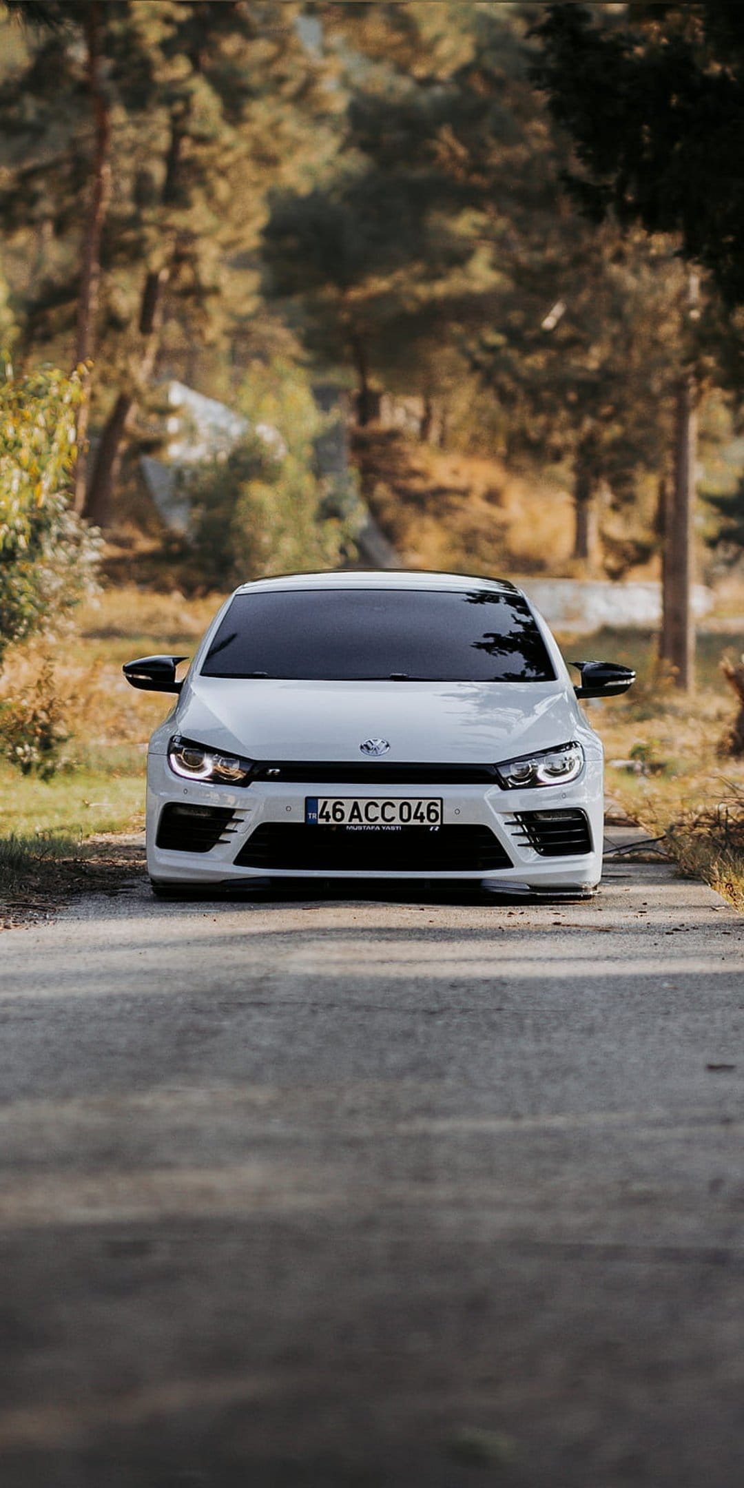 White Volkswagen Scirocco with lit headlights parked on a paved road surrounded by orange autumn forest trees.