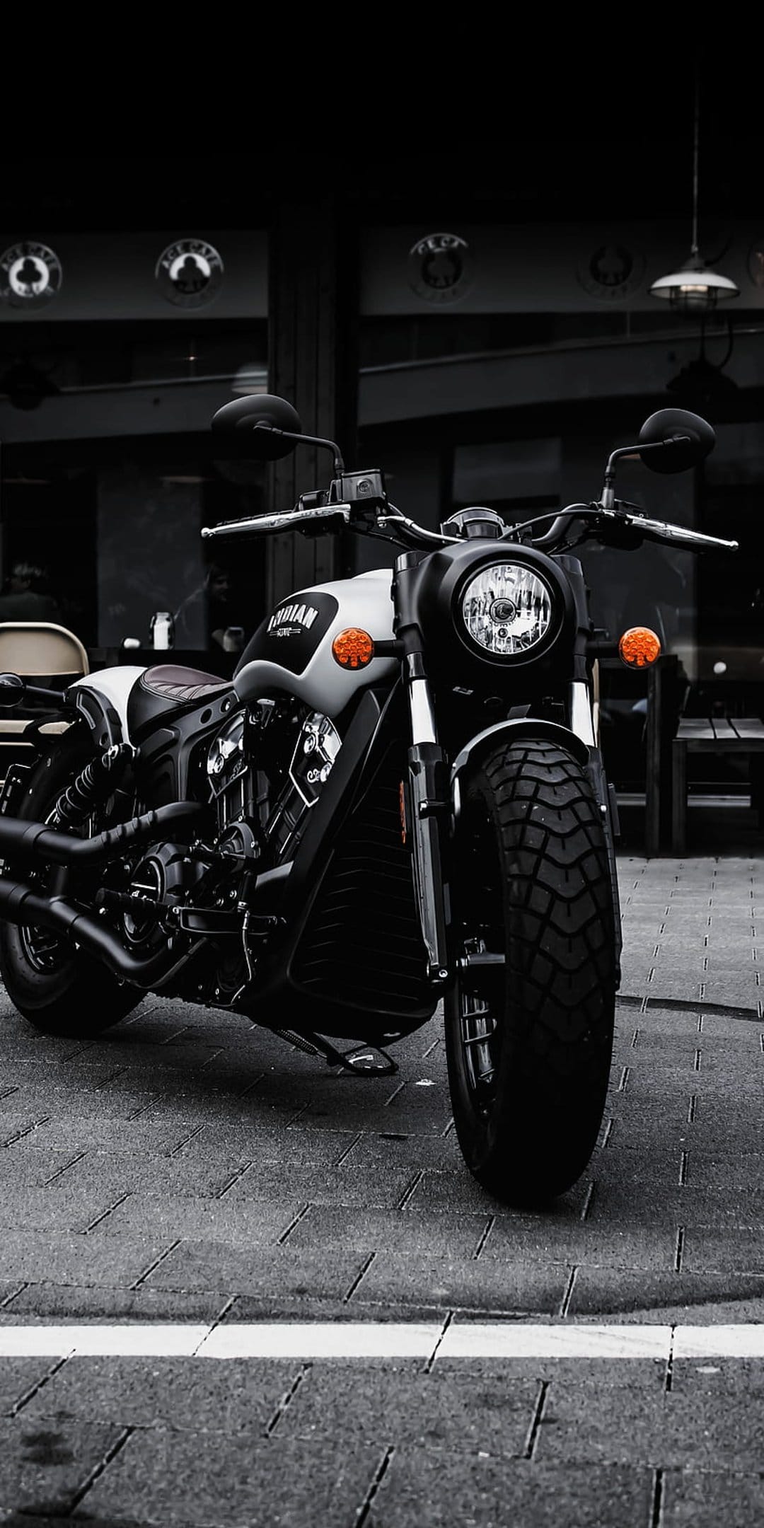 Indian motorcycle with a round headlight and dual exhaust pipes parked on a textured cobblestone city street.
