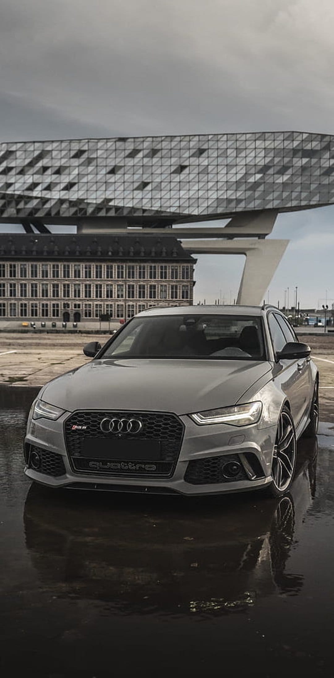 Audi RS6 Avant in gray parked on wet pavement before a faceted modern building under an overcast sky.