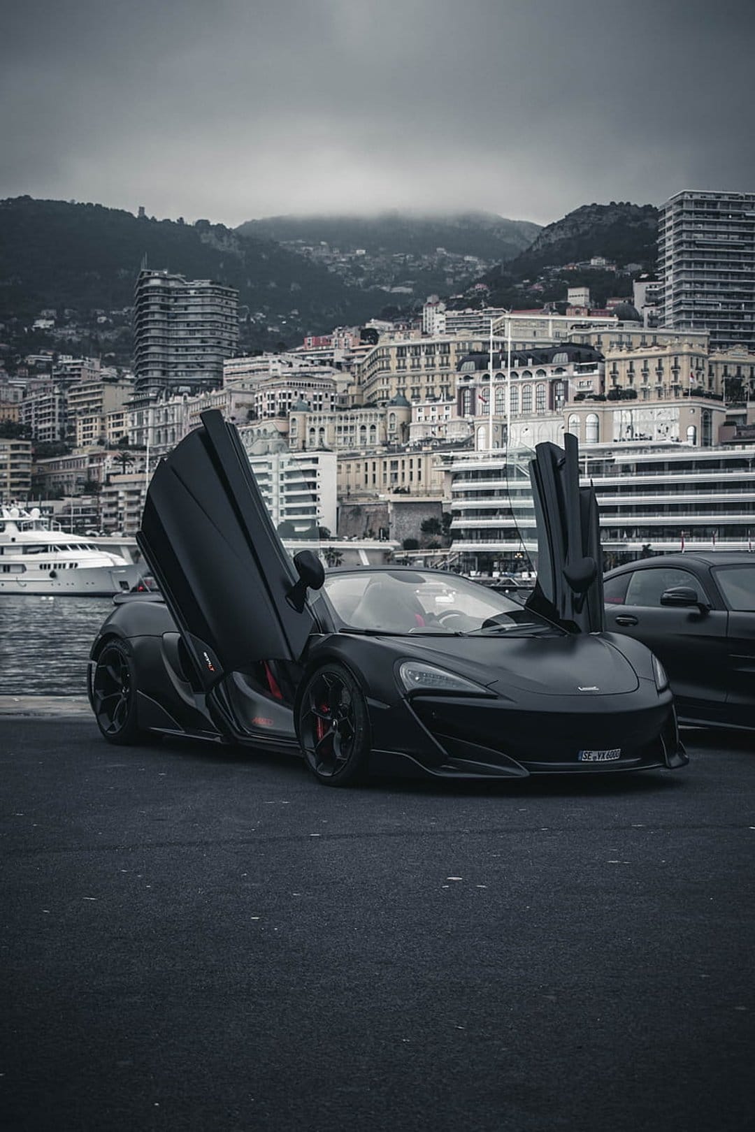 Matte black McLaren supercar with open butterfly doors parked along the Monaco harbor under a moody overcast sky.
