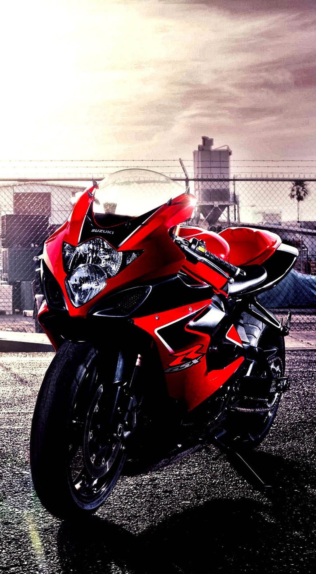 Red and black Suzuki sportbike parked on dark asphalt with an industrial urban background under a cloudy sky.
