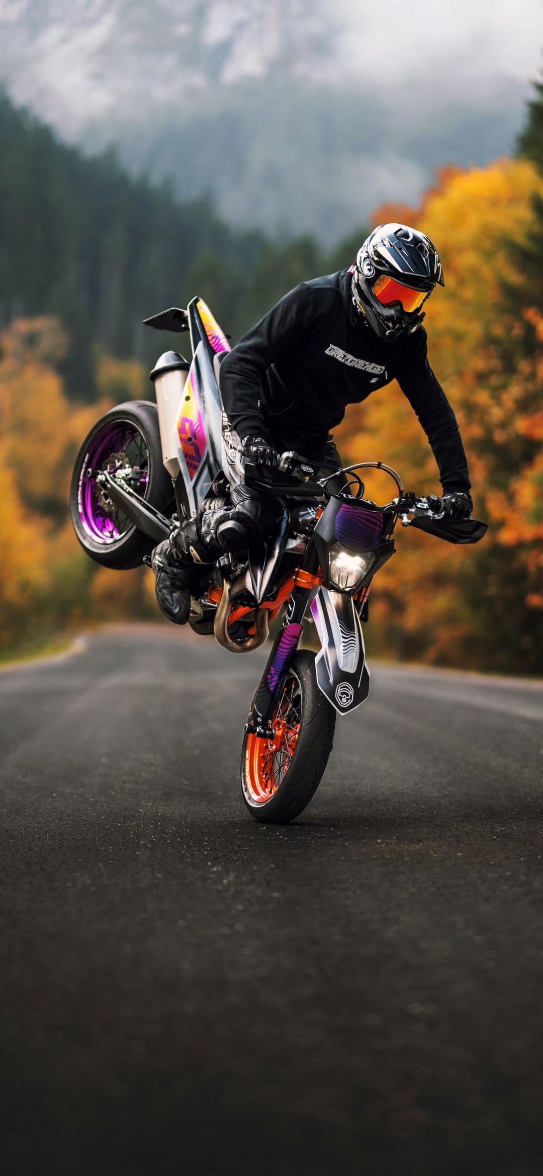 Motorcyclist in orange goggles performs a wheelie on a purple bike amidst golden autumn trees and mountains.
