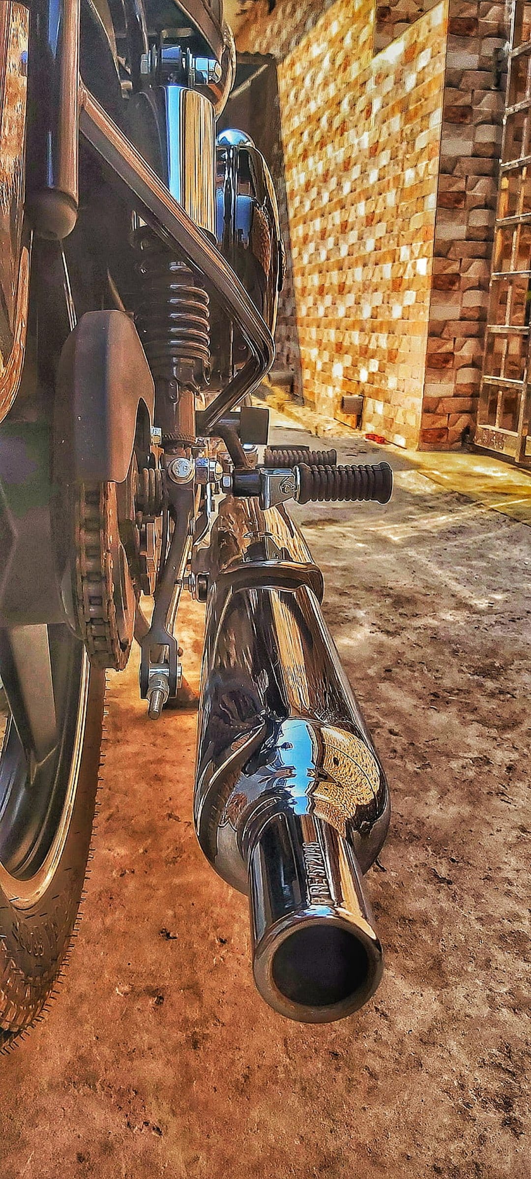 Chrome motorcycle exhaust pipe reflecting a brick wall next to a drive chain and rear suspension components.