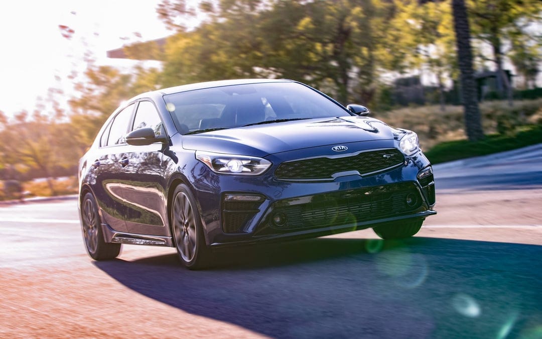 Dark blue Kia Forte GT sedan driving on a sunlit road with golden hour lens flare and motion blur.